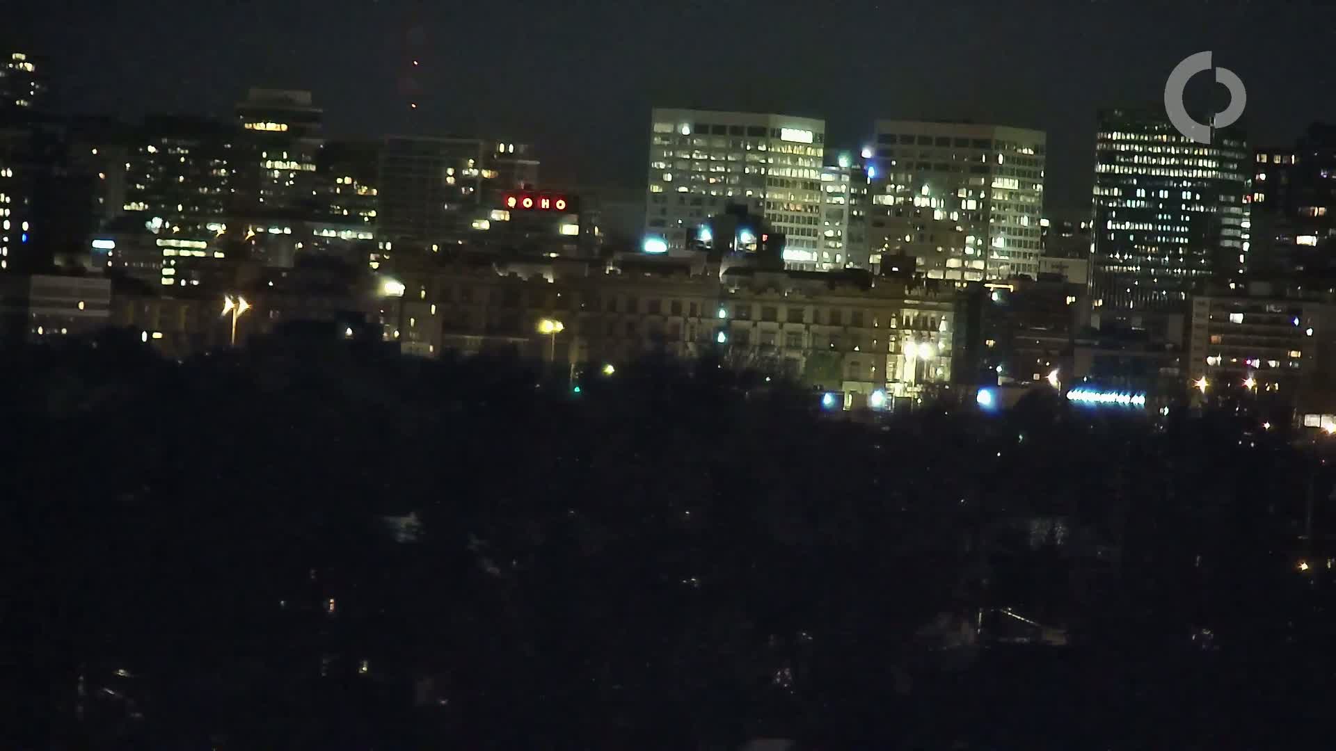 A nighttime cityscape with numerous illuminated buildings is seen from a distance, obscured partially by a dark foreground.