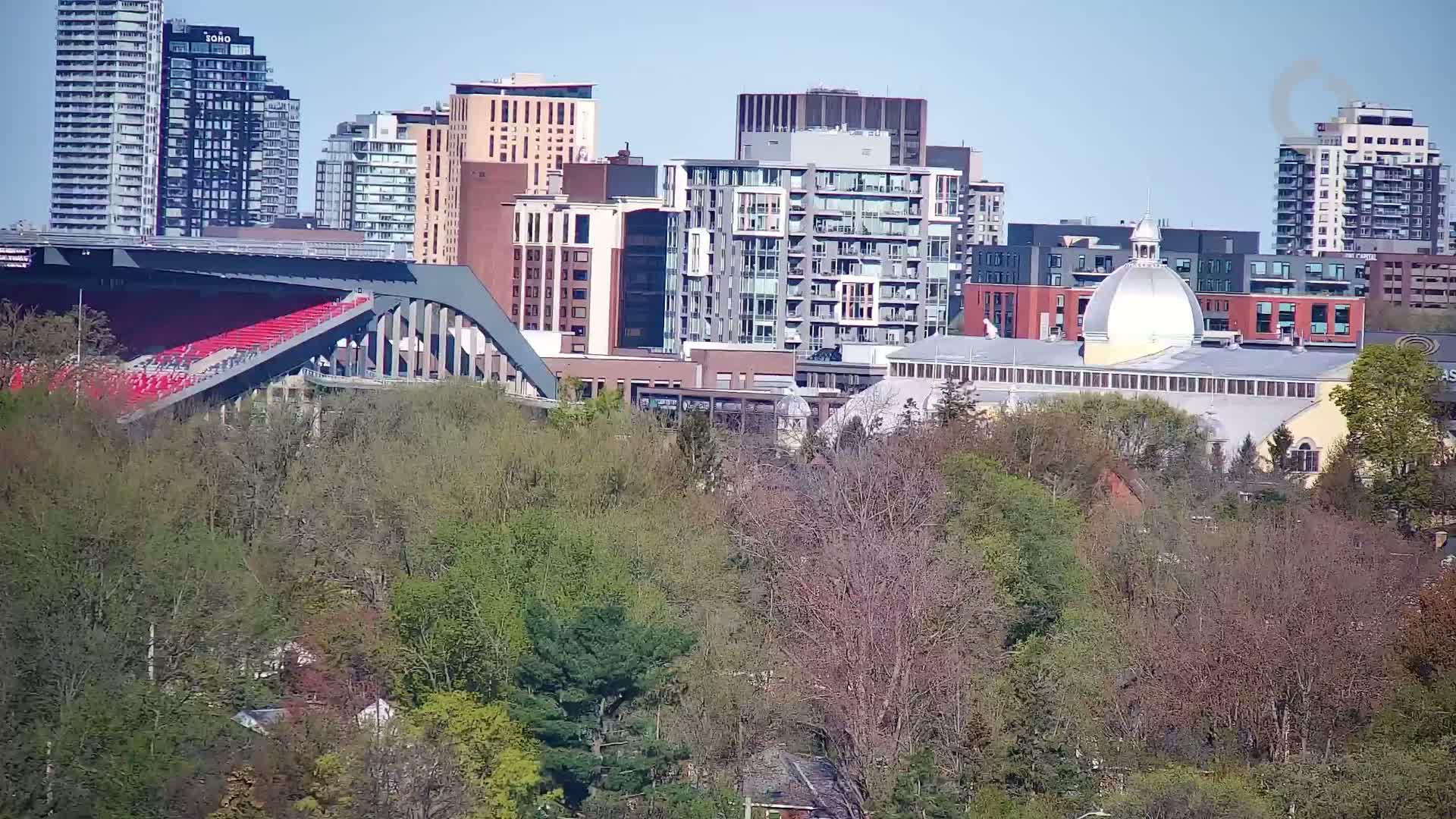 A city skyline with numerous high-rise buildings, a red stadium seating area, and a large building with a dome is partially obscured by a lush green and brown deciduous forest under a clear blue sky.