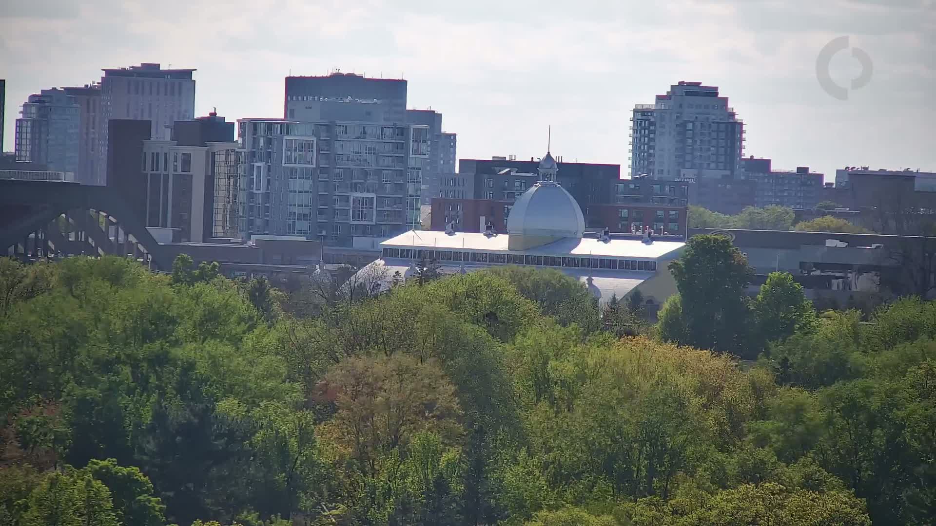 A city skyline with several multi-story buildings, a domed building partially obscured by trees, and a bridge visible in the distance under a partly cloudy sky.