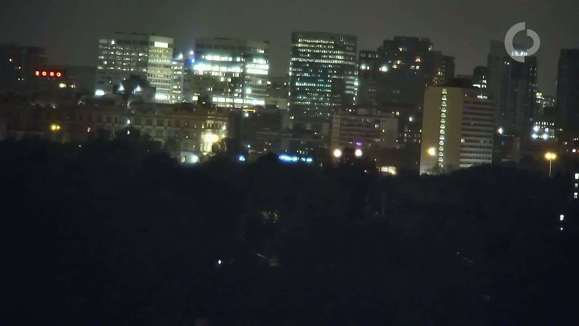 A nighttime cityscape of illuminated buildings, viewed from across a dark expanse of trees or vegetation.