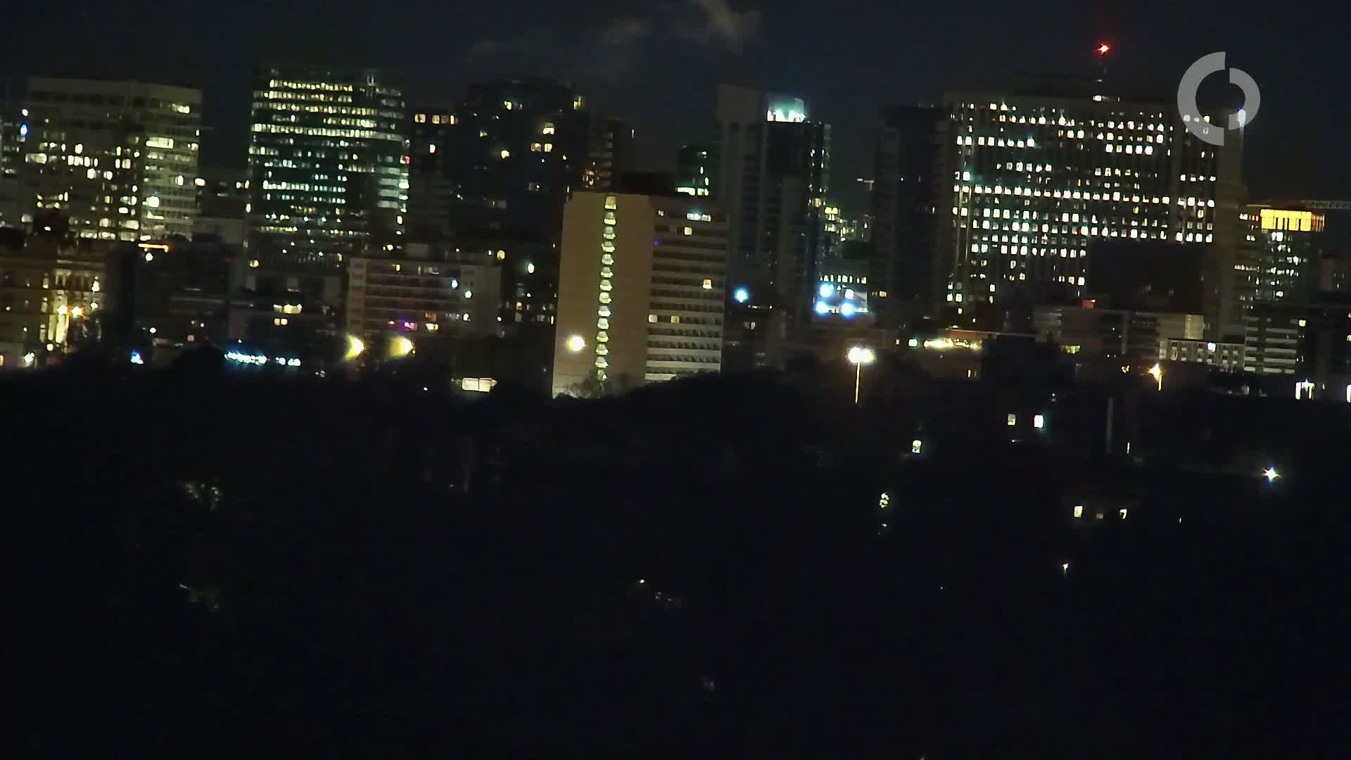 A nighttime view of a city skyline with numerous illuminated buildings, viewed from a distance across a dark foreground.