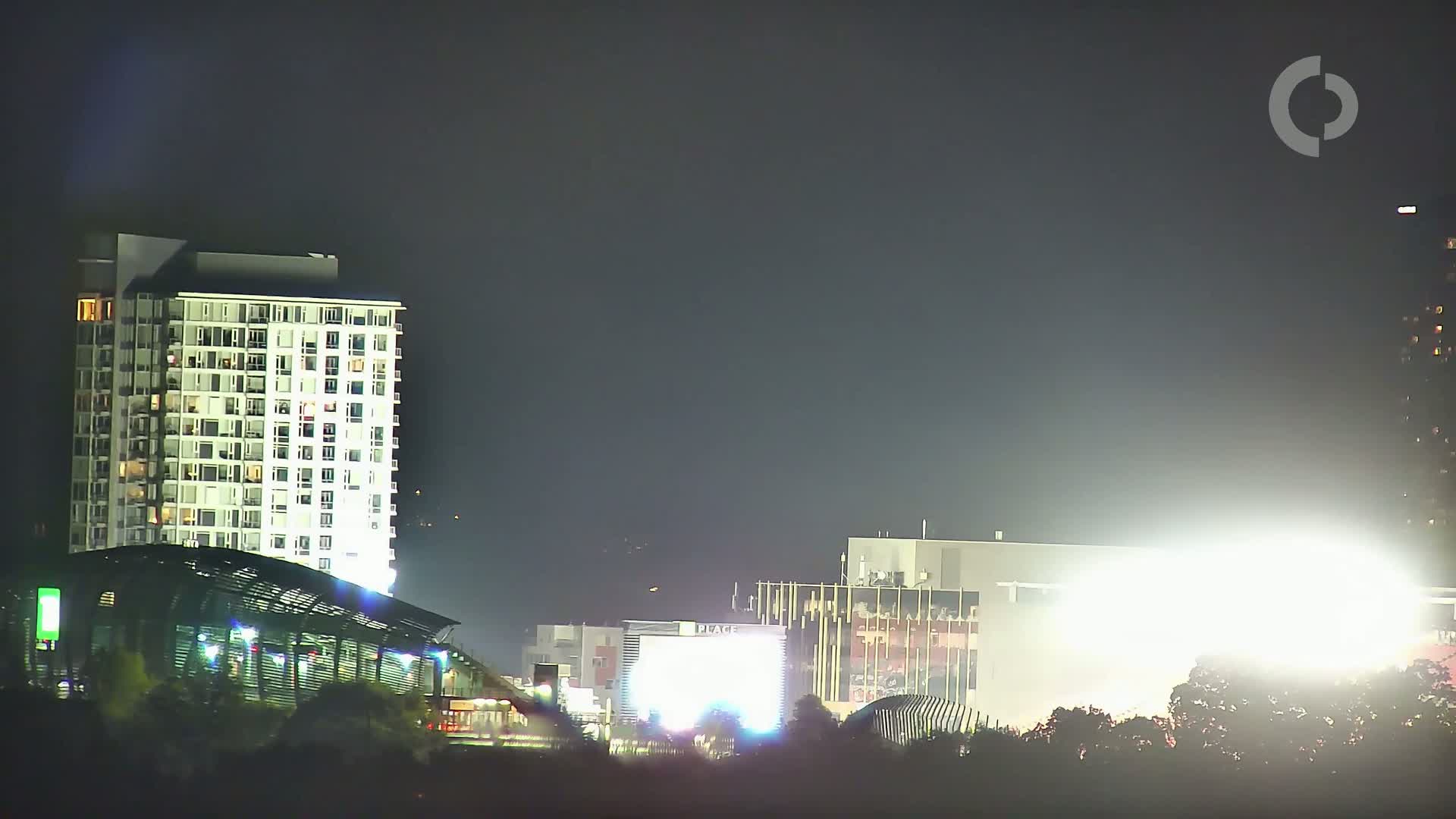 A brightly lit city skyline at night, featuring a tall apartment building and other structures partially obscured by trees and bright lights.