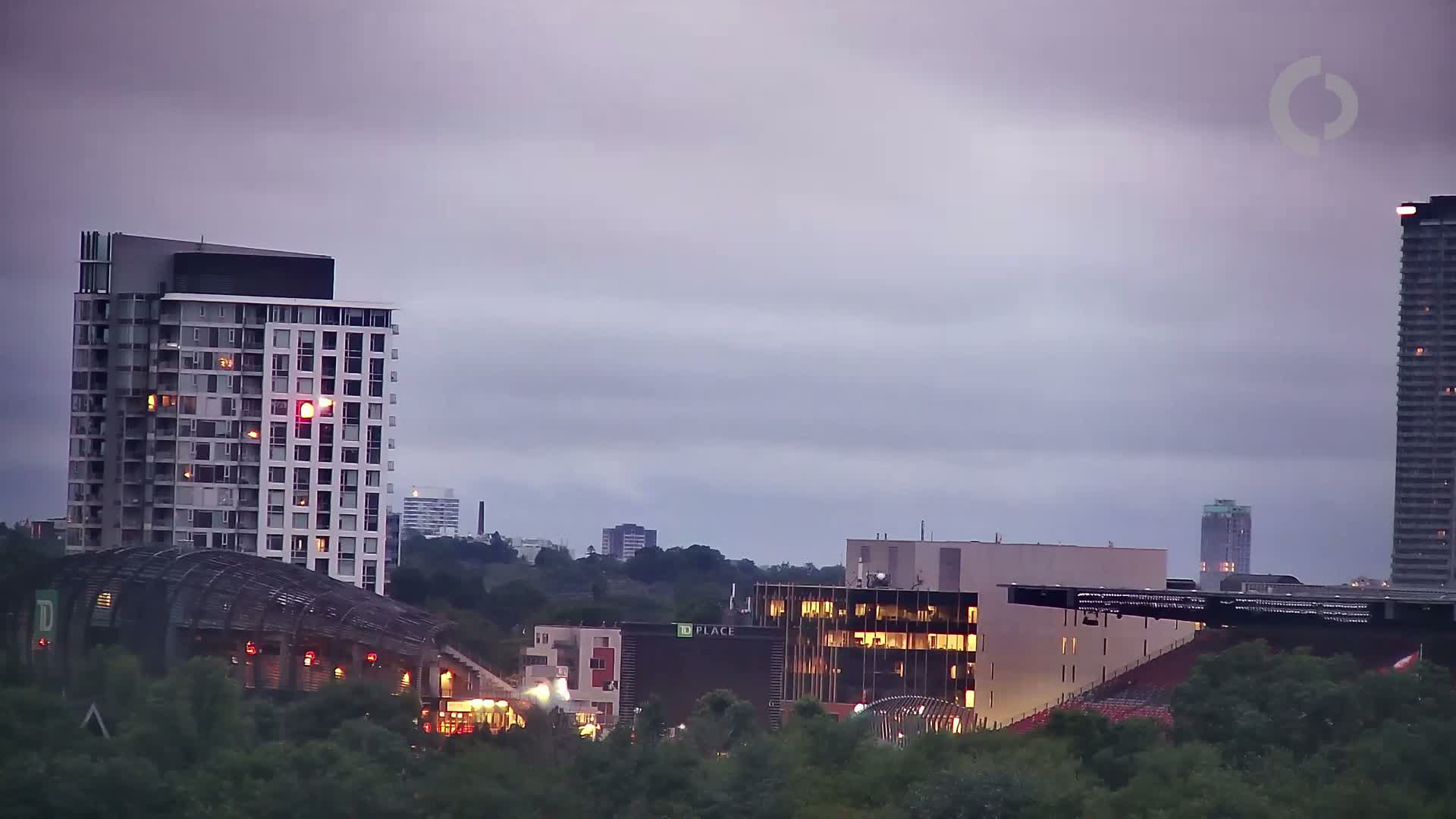 A city skyline at dusk under a cloudy sky features several high-rise buildings, some with illuminated windows, and a partially visible stadium.