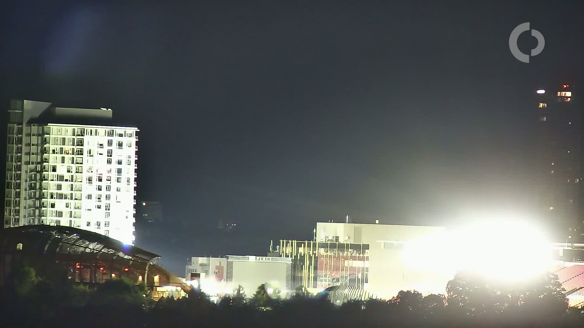 At night, brightly lit buildings, including a tall apartment building and other structures, are visible under a dark sky.
