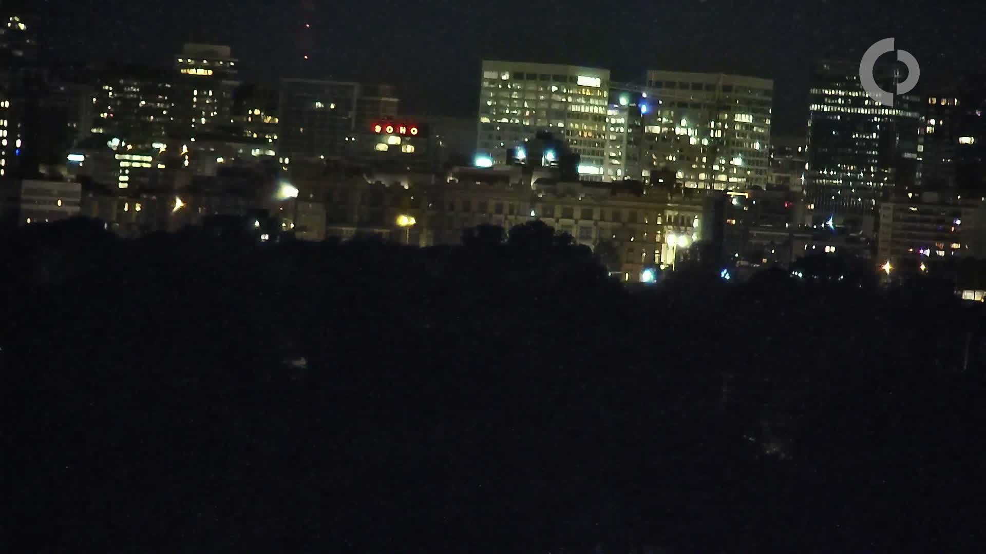A nighttime cityscape with numerous buildings illuminated, viewed from a distance across a dark foreground.