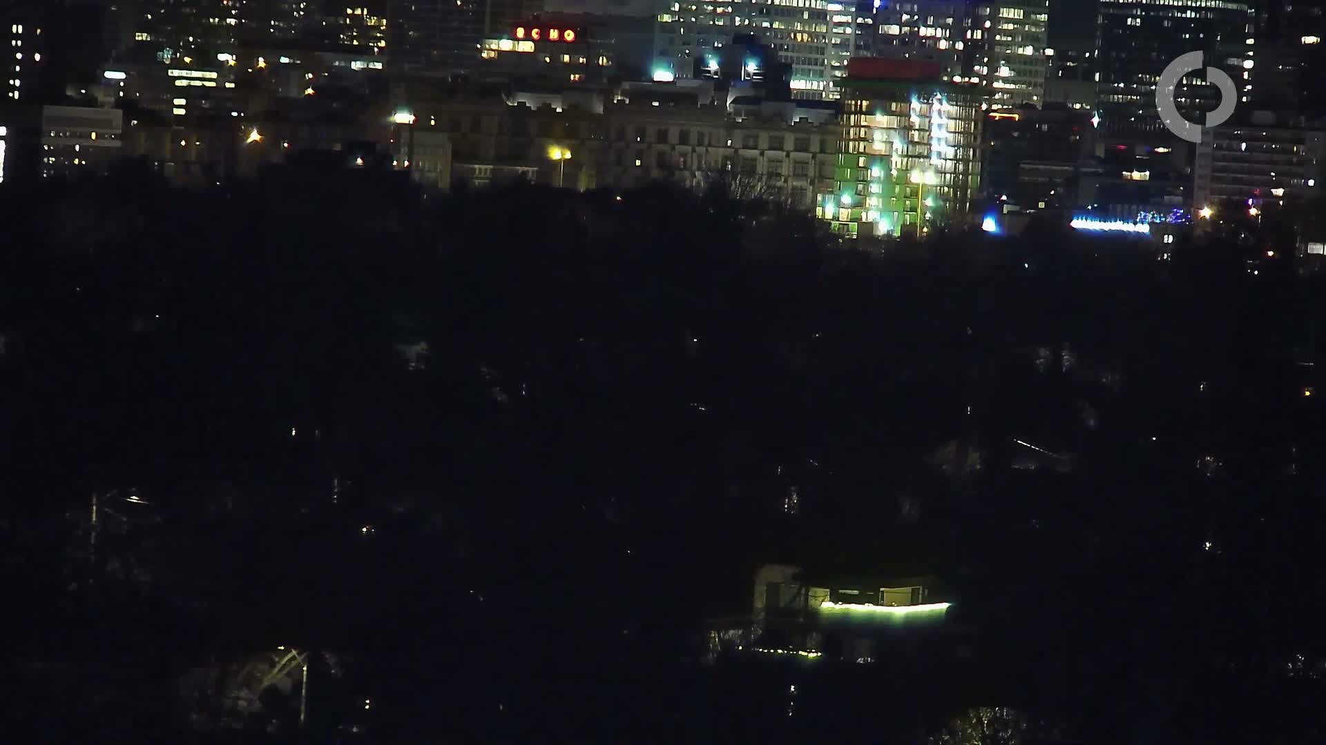 A nighttime aerial view reveals a sprawling city skyline illuminated by countless lights in the background, contrasting with a dark, silhouetted landscape featuring scattered distant lights in the foreground, all under clear weather conditions.