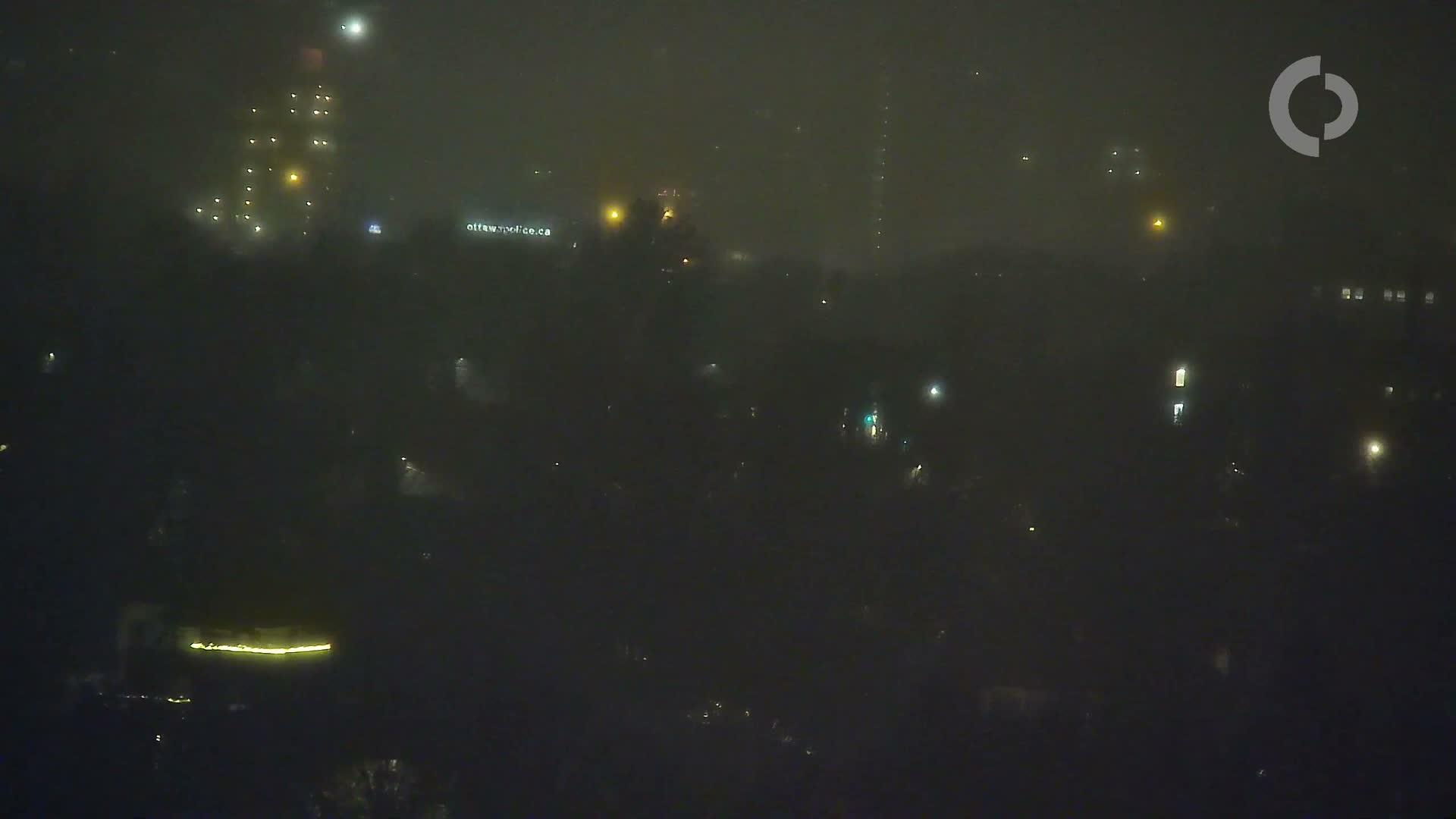 A nighttime city scene is enveloped in heavy fog, with building and street lights appearing as hazy, diffused glows amidst the low visibility.