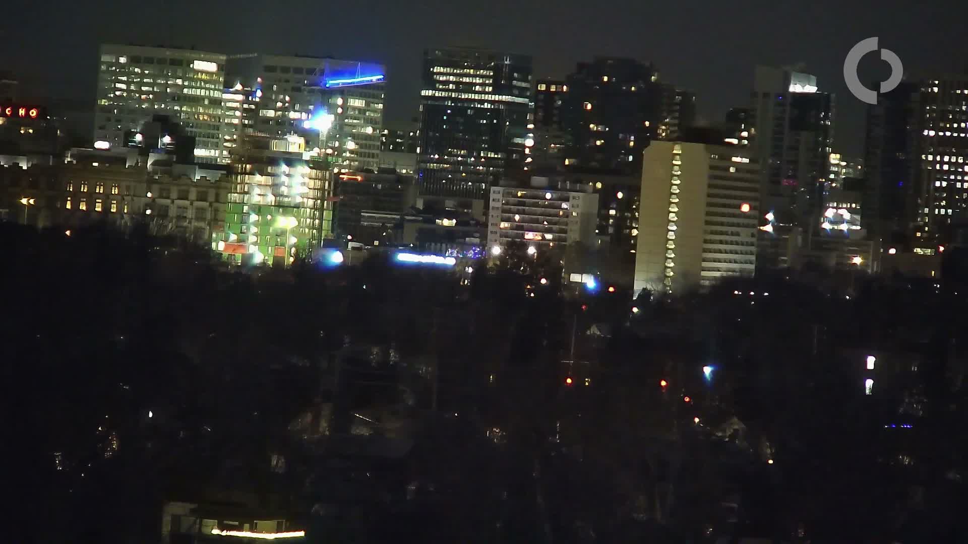 A distant city skyline glows brightly at night under a clear dark sky, with numerous illuminated buildings visible above a dark, presumably treed, foreground.