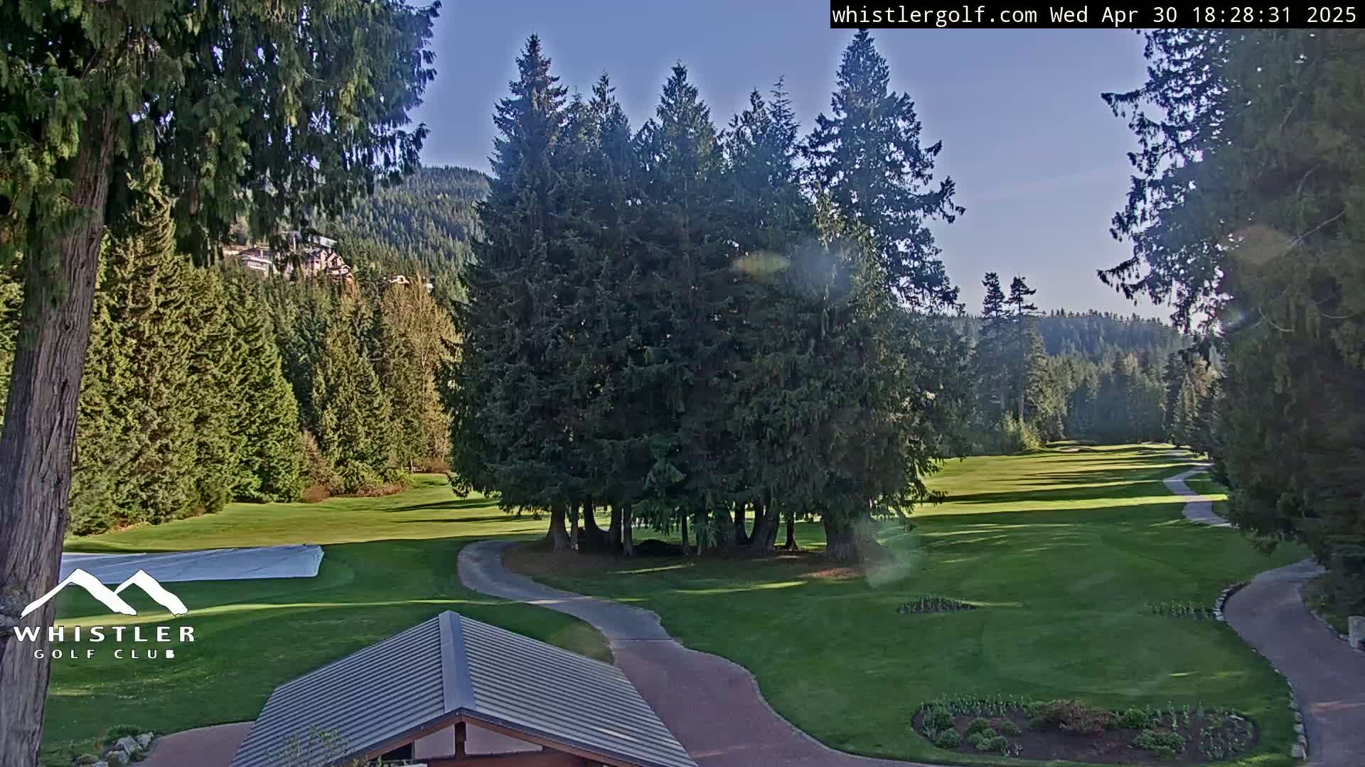 A sunny day reveals a golf course nestled among evergreen trees and mountains, with a paved pathway winding through the green fairways.