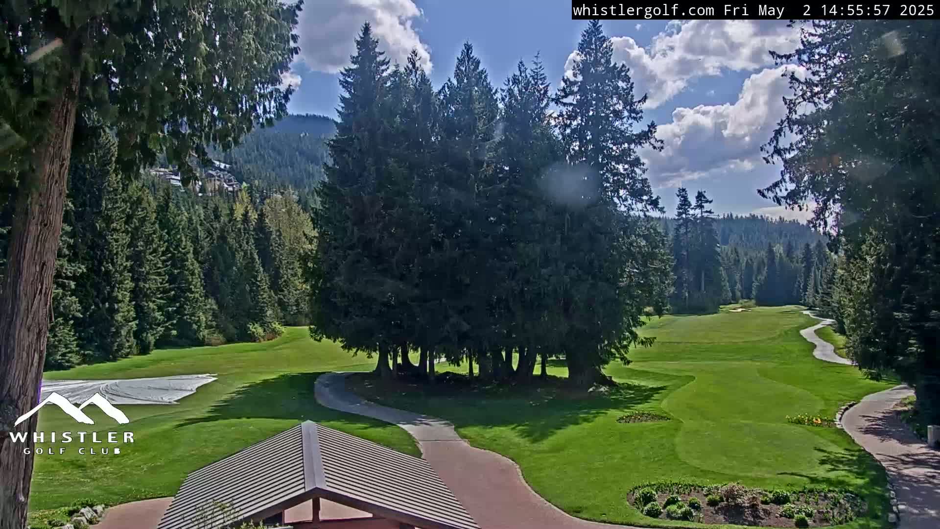 A partly sunny day reveals a green golf course nestled among evergreen trees and mountains.