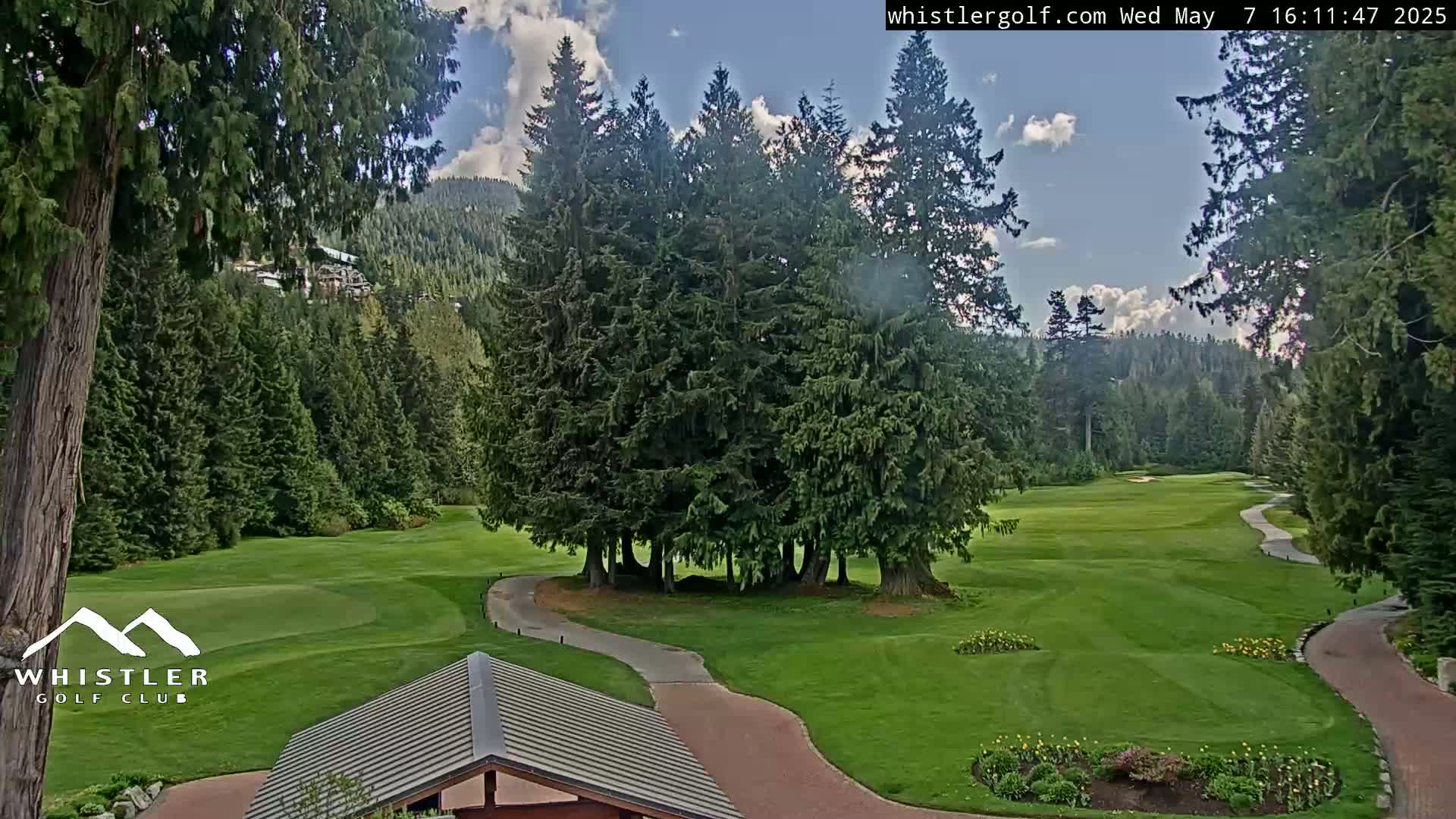 A partly cloudy sky overlooks a green golf course surrounded by lush evergreen trees and winding pathways.