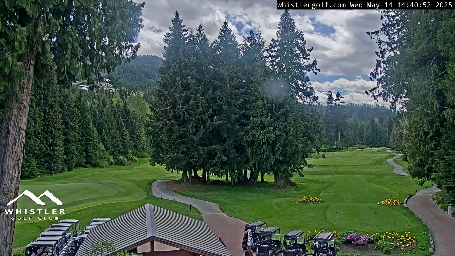 A partly cloudy day on a lush green golf course surrounded by tall evergreen trees and mountains in the distance.