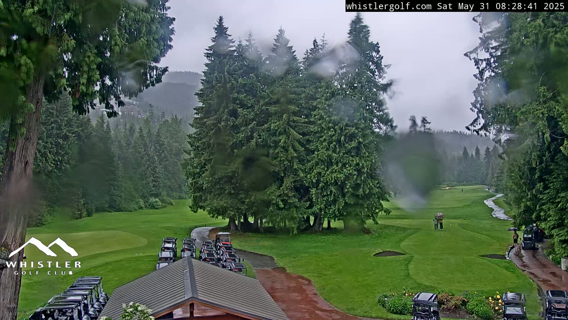 A rain-soaked golf course, nestled among tall evergreen trees and mountains, shows several golf carts parked near a clubhouse with a few golfers visible on the course.