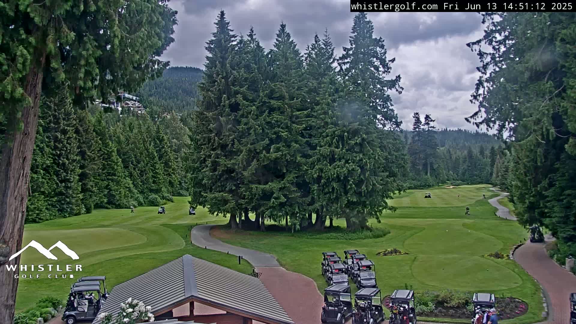 A golf course nestled in a forest of tall evergreen trees under a partly cloudy sky, with several golf carts parked near a building.
