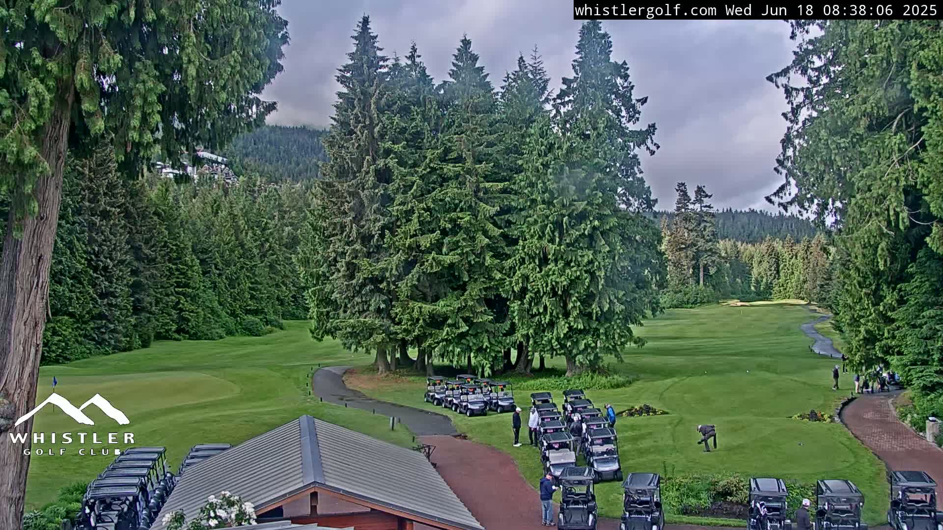 A golf course with numerous golf carts parked near a clubhouse, surrounded by lush evergreen trees under a partly cloudy sky.