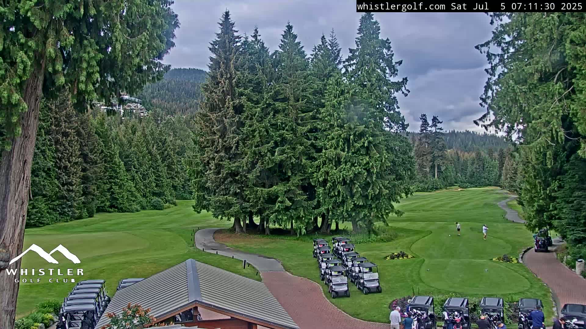 A golf course nestled in a forest of tall evergreen trees under a cloudy sky, with golf carts parked near a clubhouse and golfers on the green.