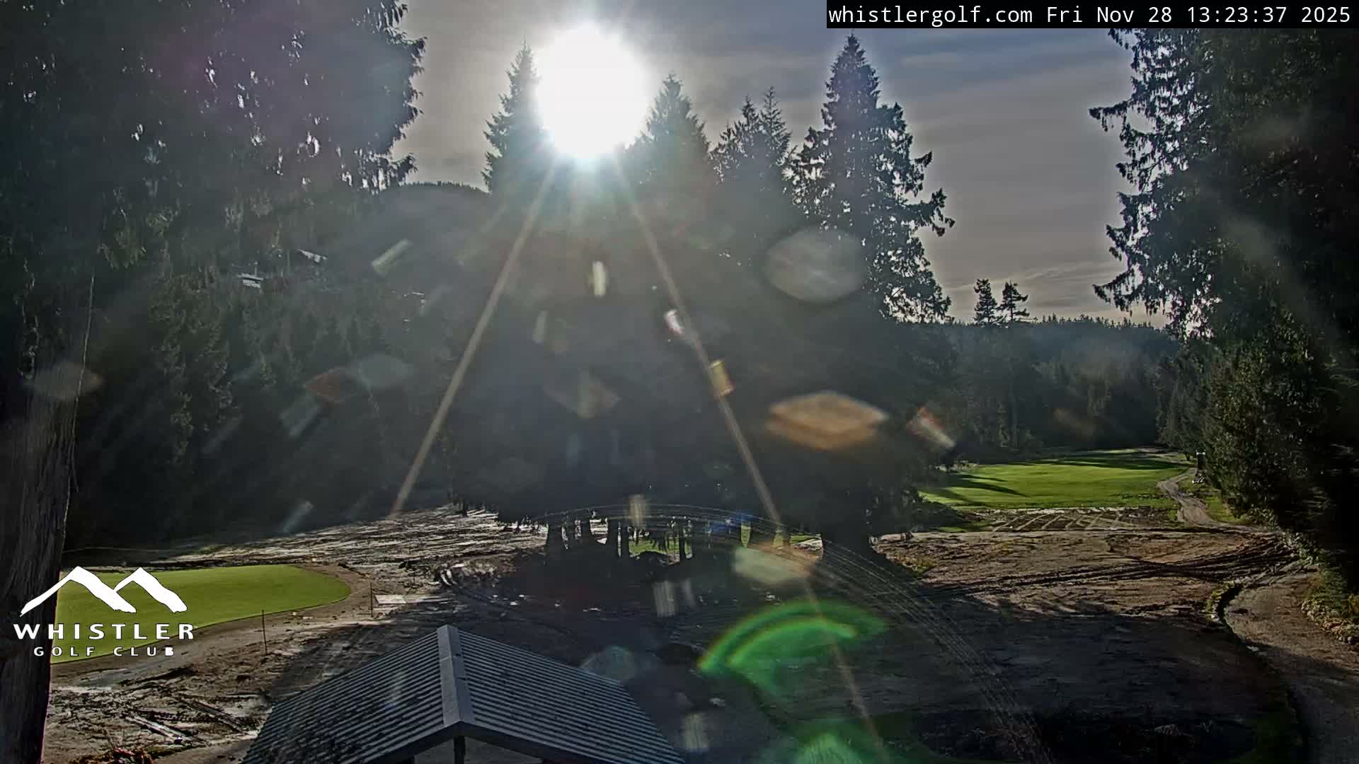 A golf course is seen under bright, sunny, partly cloudy skies, featuring verdant greens, bare earth pathways, a small bridge, and surrounded by dense evergreen forests.