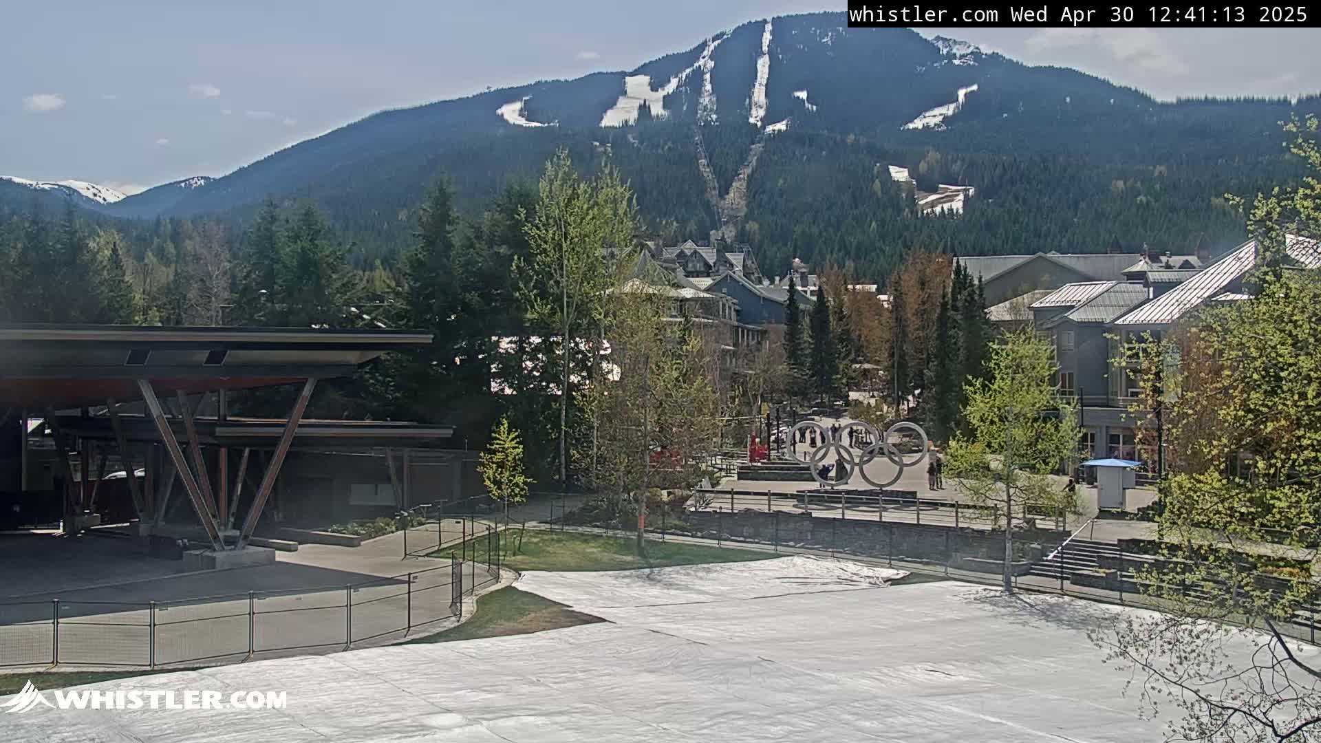 A sunny mountain village scene features a large snow-covered area in the foreground, a modern building to the left, and a mountain with ski runs in the background.
