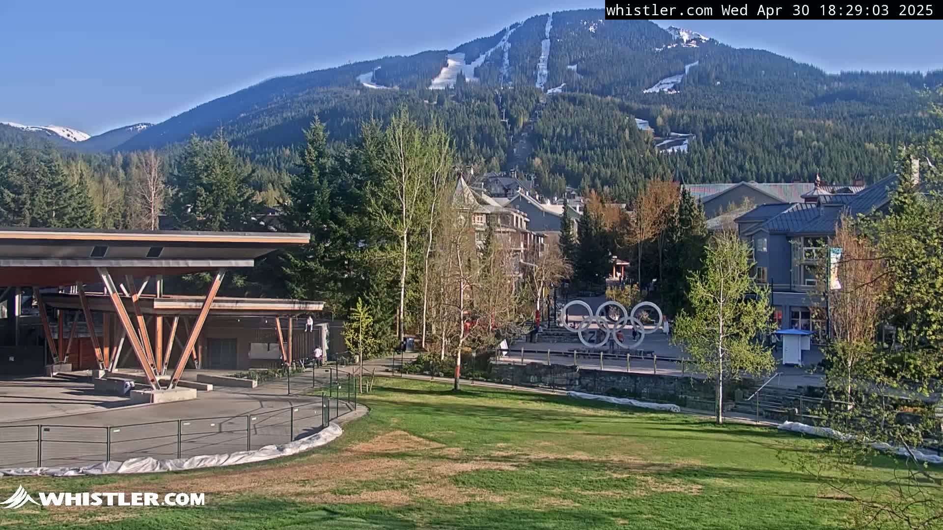 A sunny view of a village nestled in a mountain valley with ski slopes visible on the mountainside, featuring a grassy area with a modern structure in the foreground.