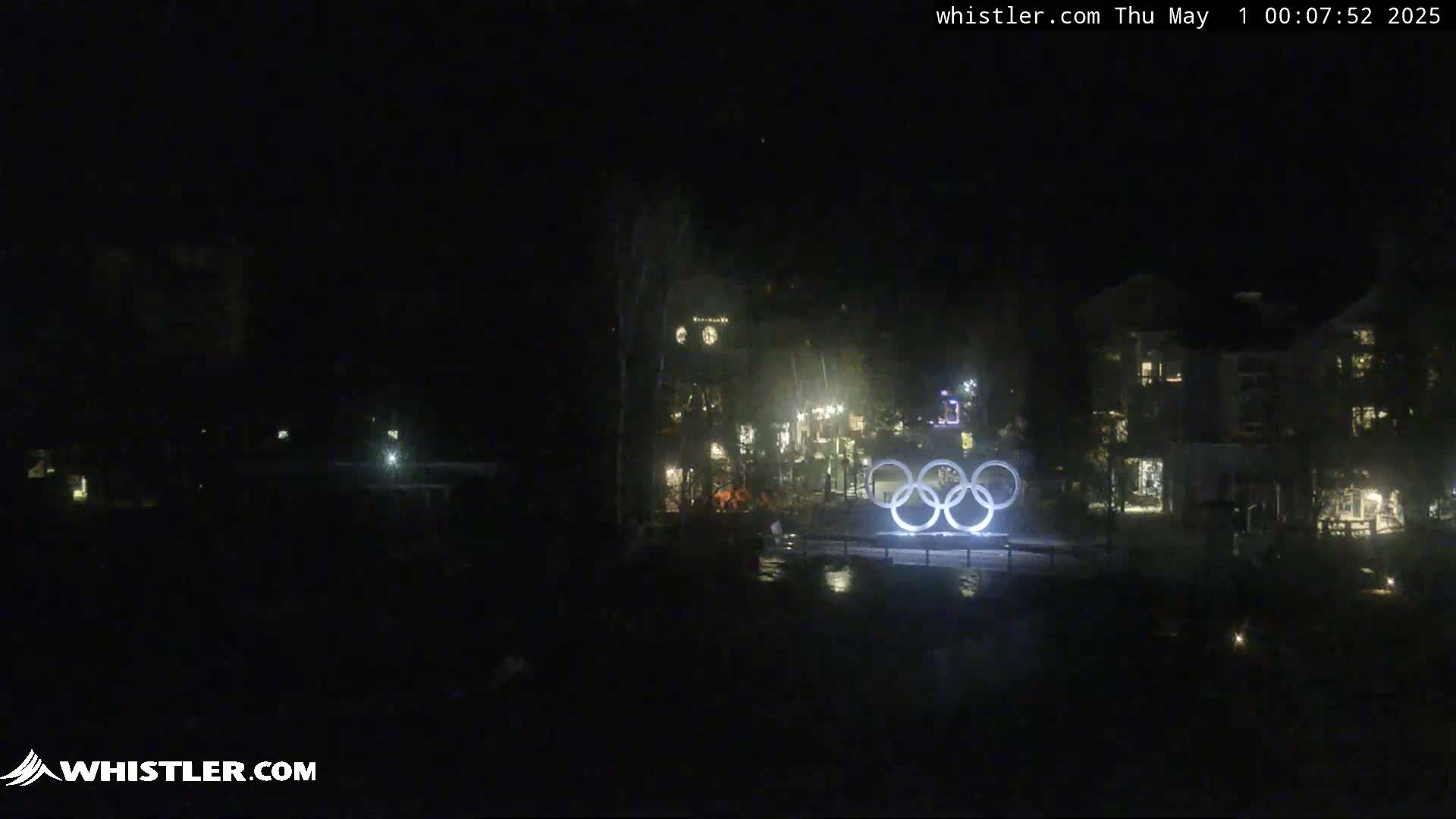 At night, illuminated Olympic rings stand in front of a dark building complex under a clear, dark sky.