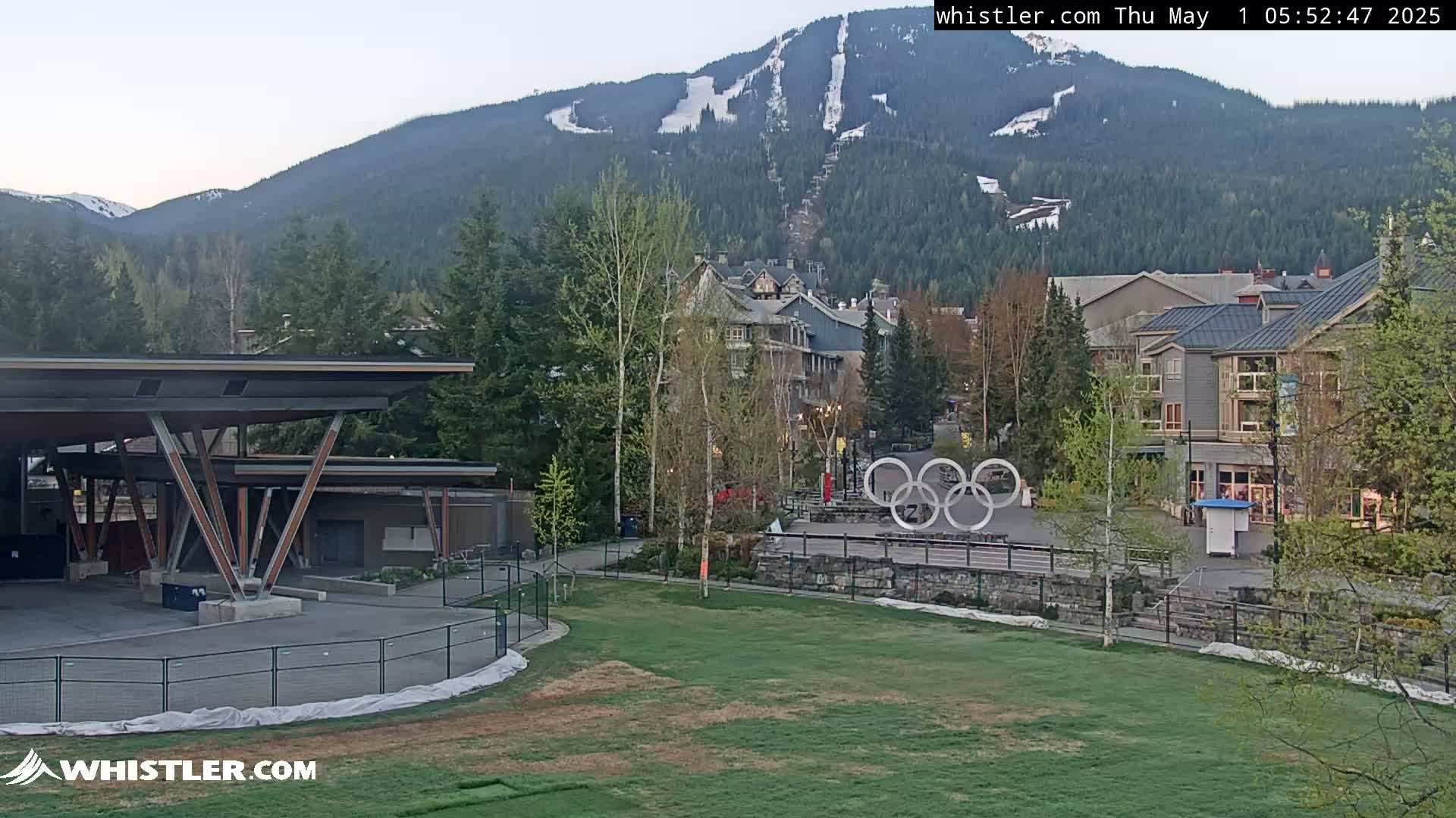 A mountainside with a ski run is visible in the background of a grassy area with buildings, trees, and the Olympic rings in the foreground, under a mostly clear sky.