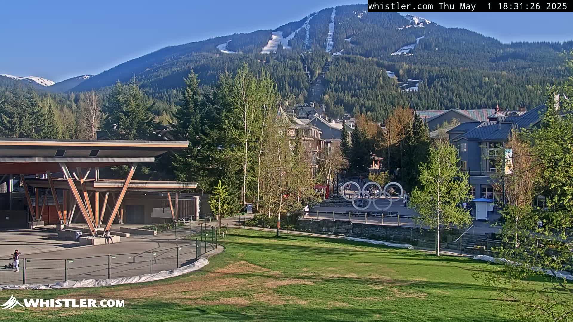 A grassy park with a modern pavilion sits in front of a village nestled at the base of a snow-capped mountain range under a clear blue sky.
