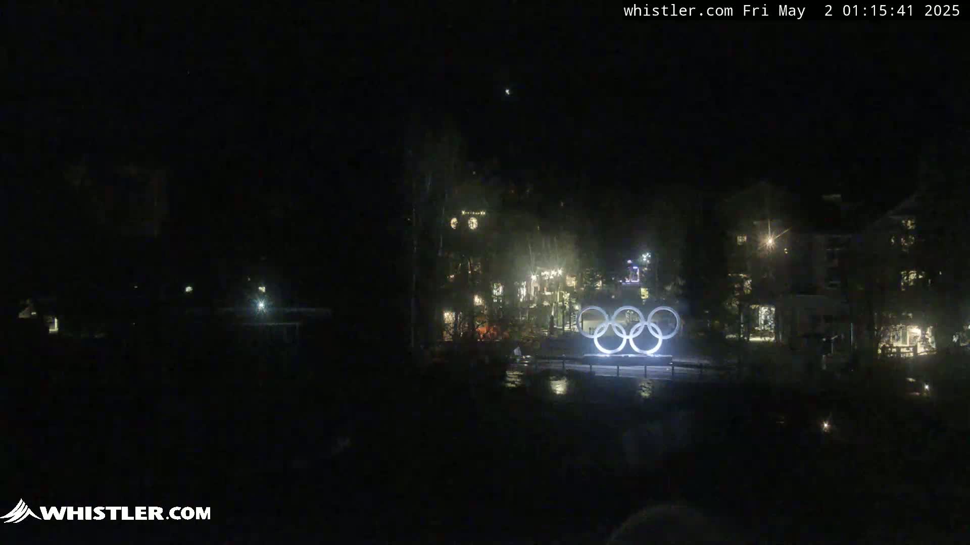At night, illuminated Olympic rings stand in front of a dark, sparsely lit village.
