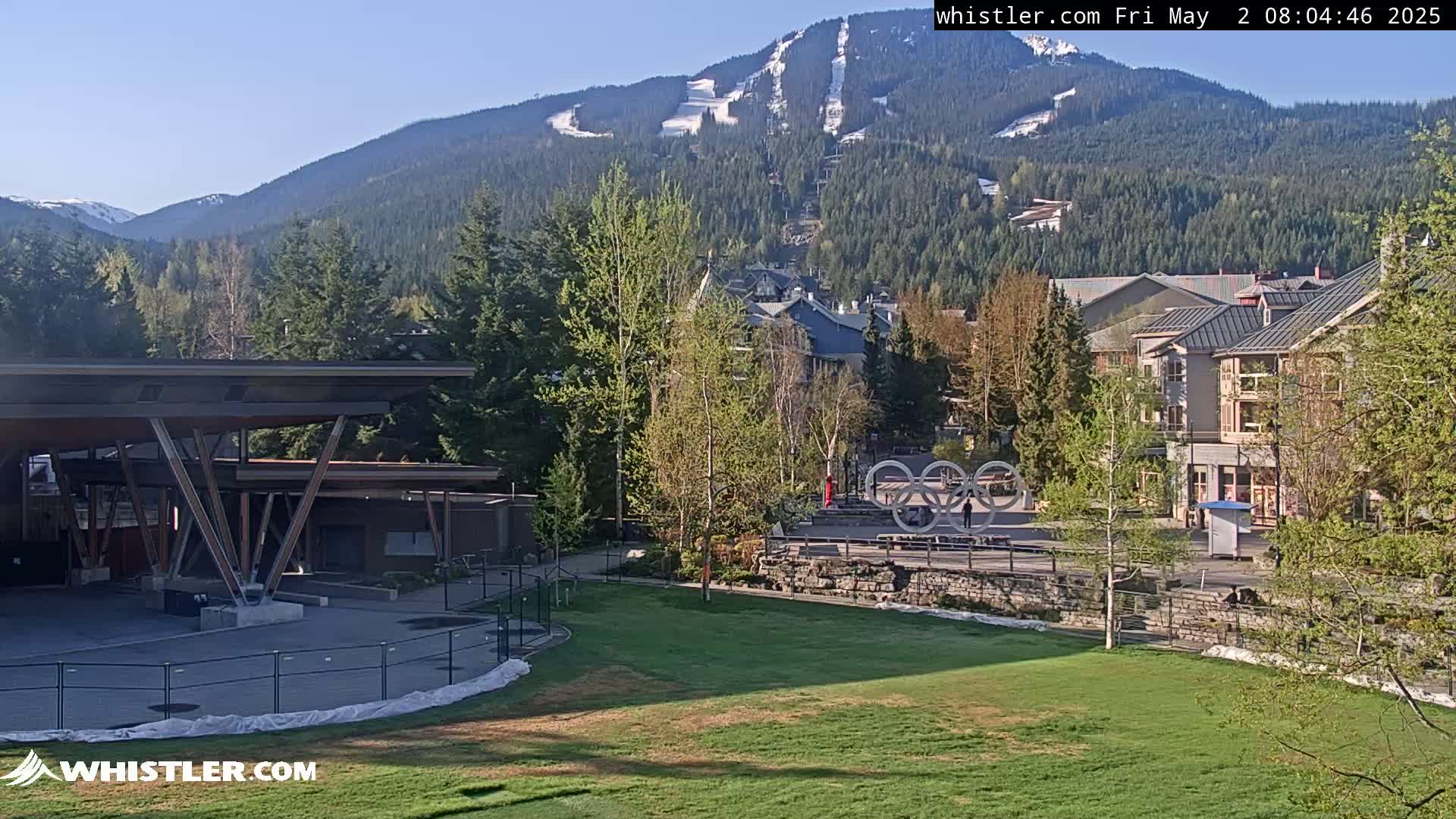 A mountain village scene on a sunny day shows a grassy field, buildings, trees in spring leaf, and a mountain range in the background with a ski run visible.