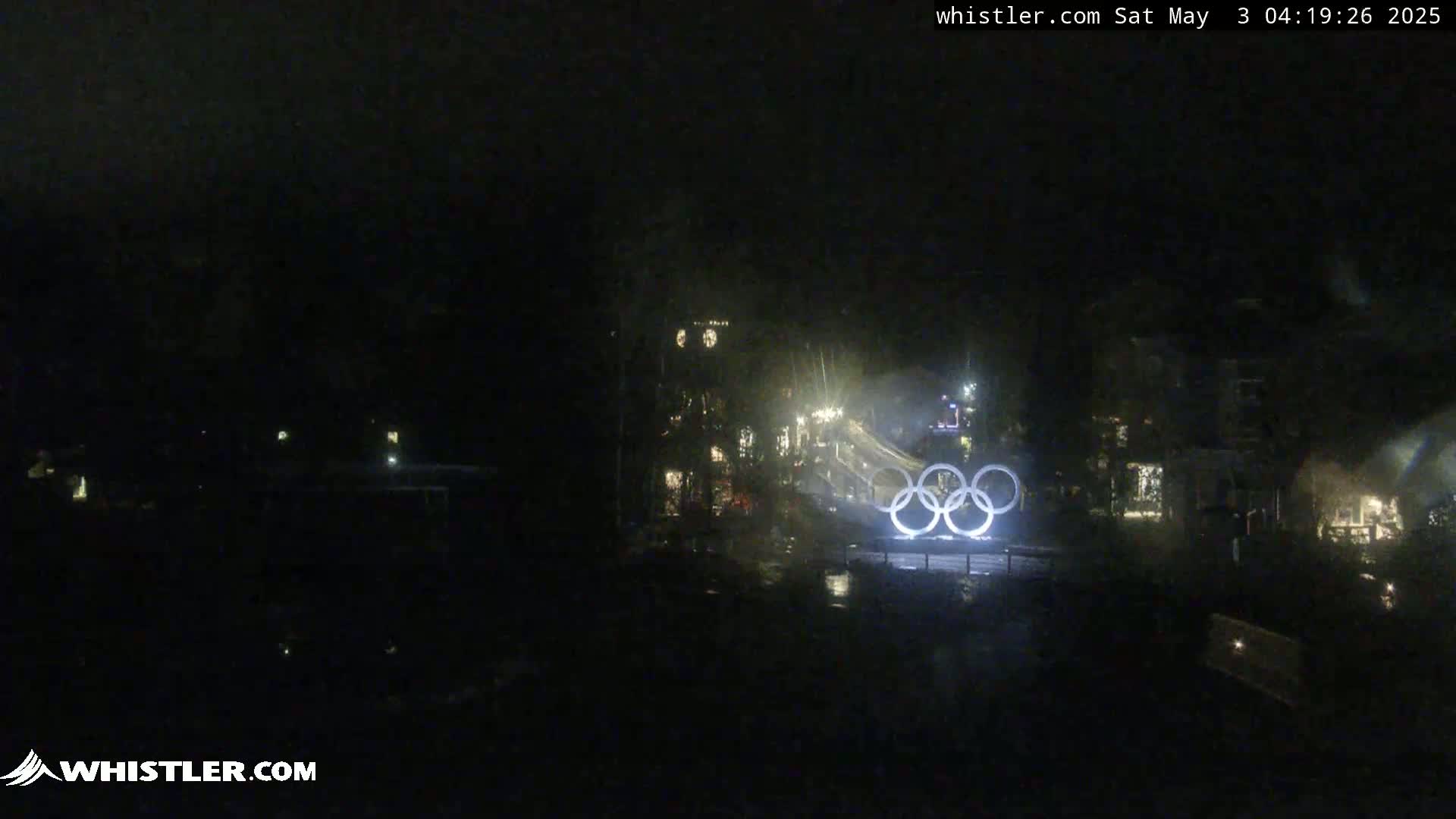 At night, illuminated Olympic rings are visible in the distance, with a dark, possibly rainy or overcast sky above a dimly lit village or town.