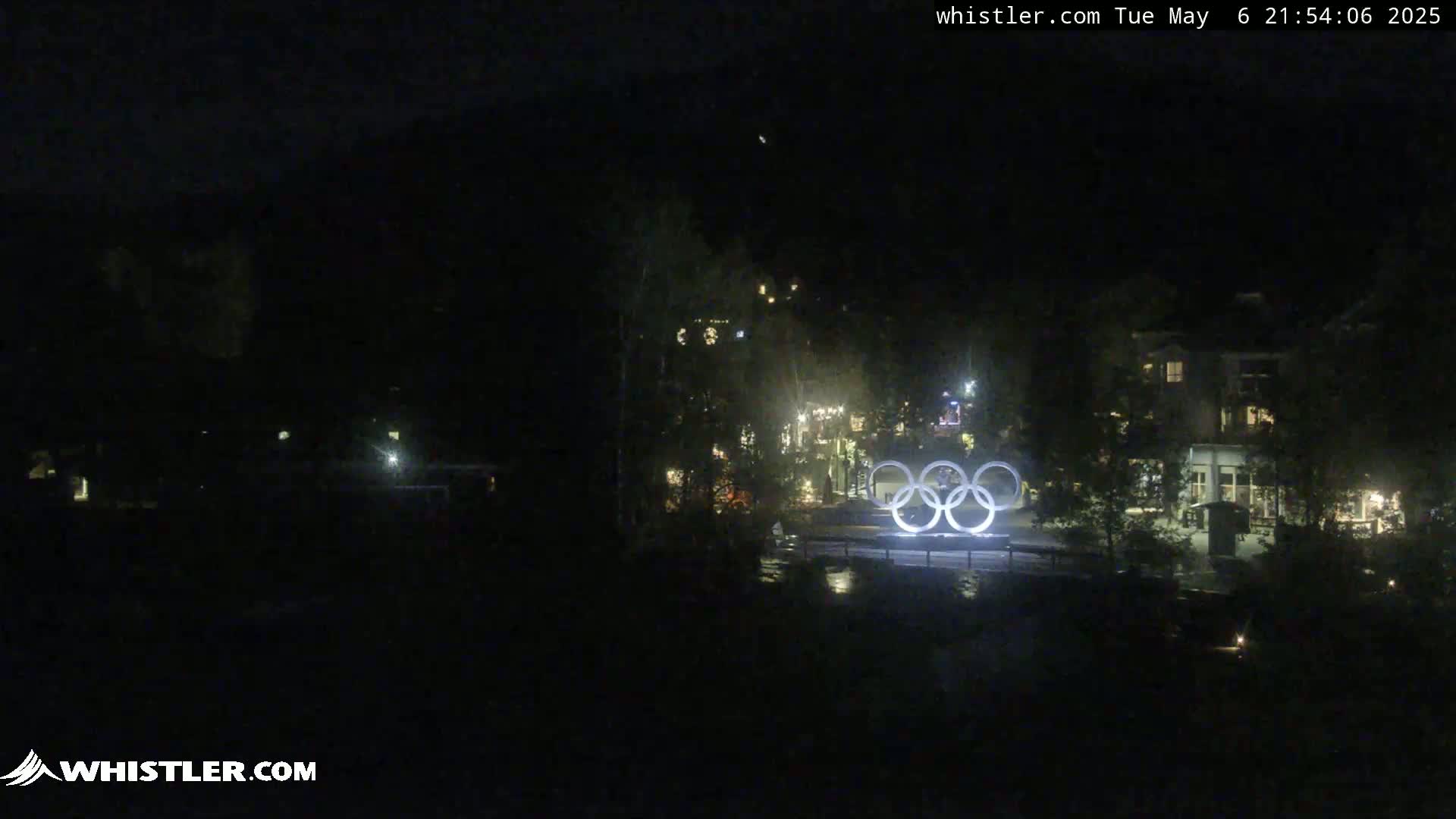 At night, illuminated Olympic rings stand in front of a dark, sparsely lit village.