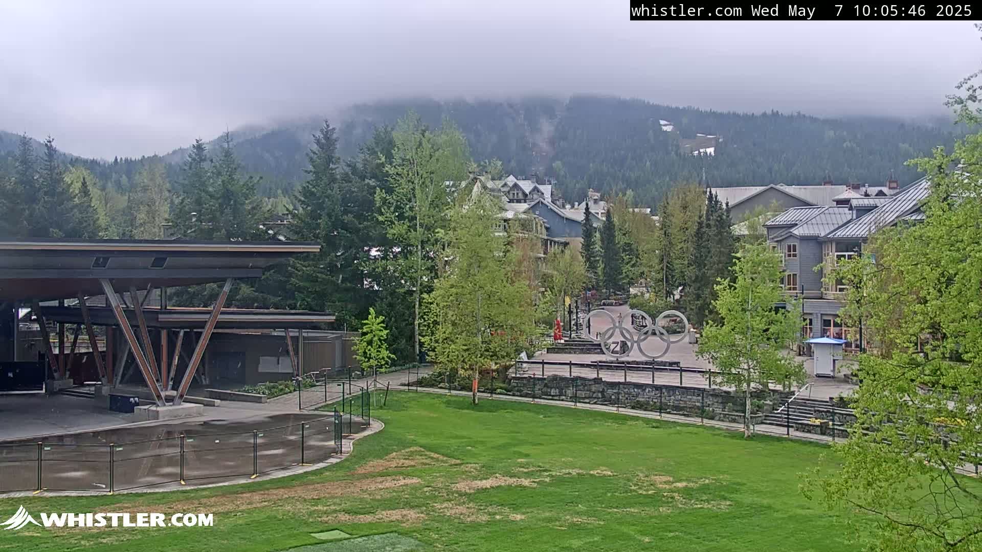 A misty mountain backdrop overlooks a grassy area with a modern structure, buildings, and the Olympic rings in a village setting.
