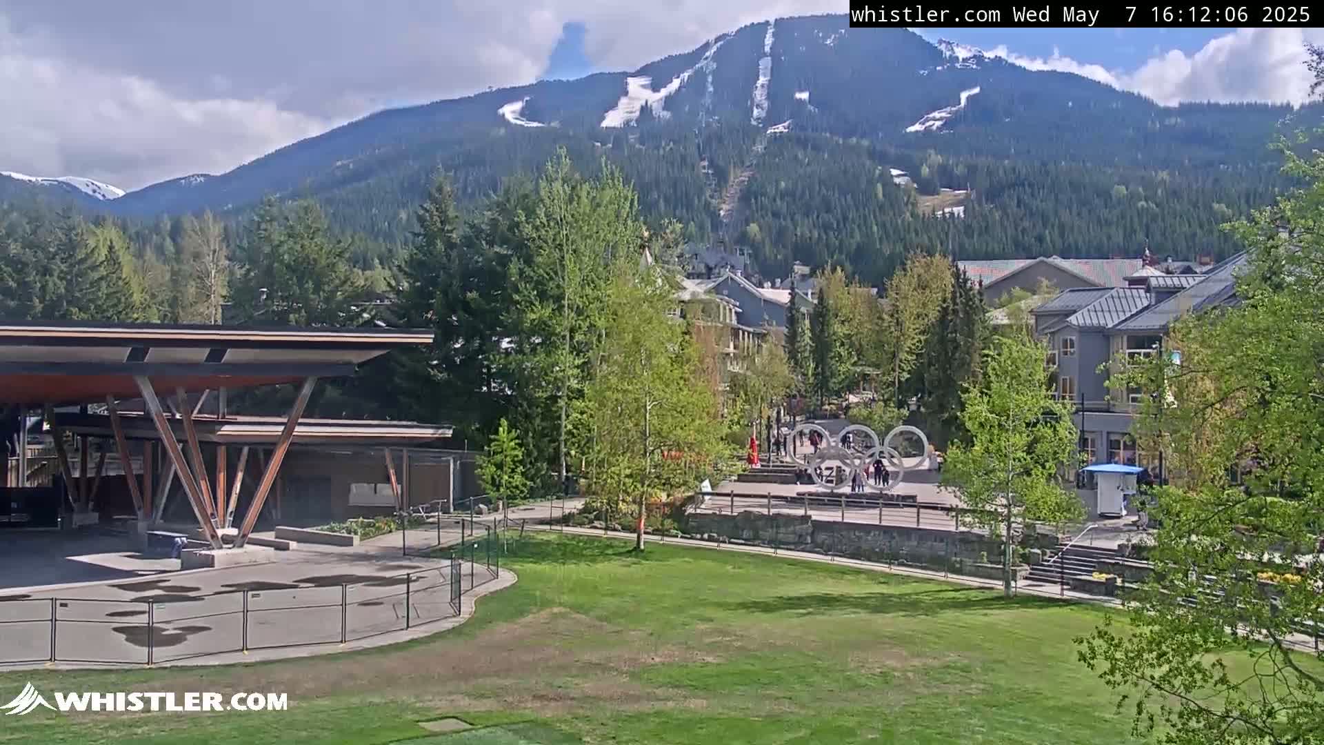 A partly sunny view of a grassy area with a modern pavilion in the foreground, overlooking a village nestled against a mountain range with visible ski runs and some remaining snow.