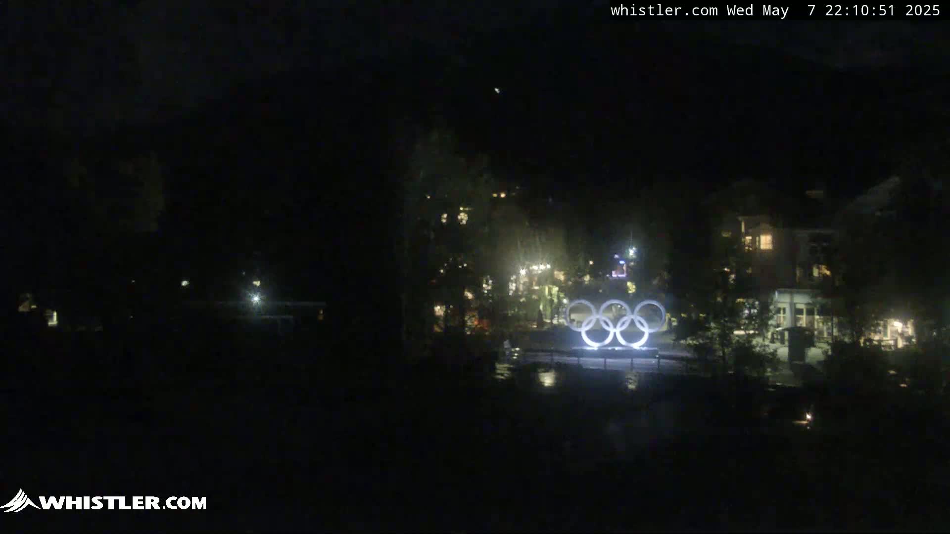 A nighttime long shot shows the illuminated Olympic rings near some buildings and trees under a dark sky.