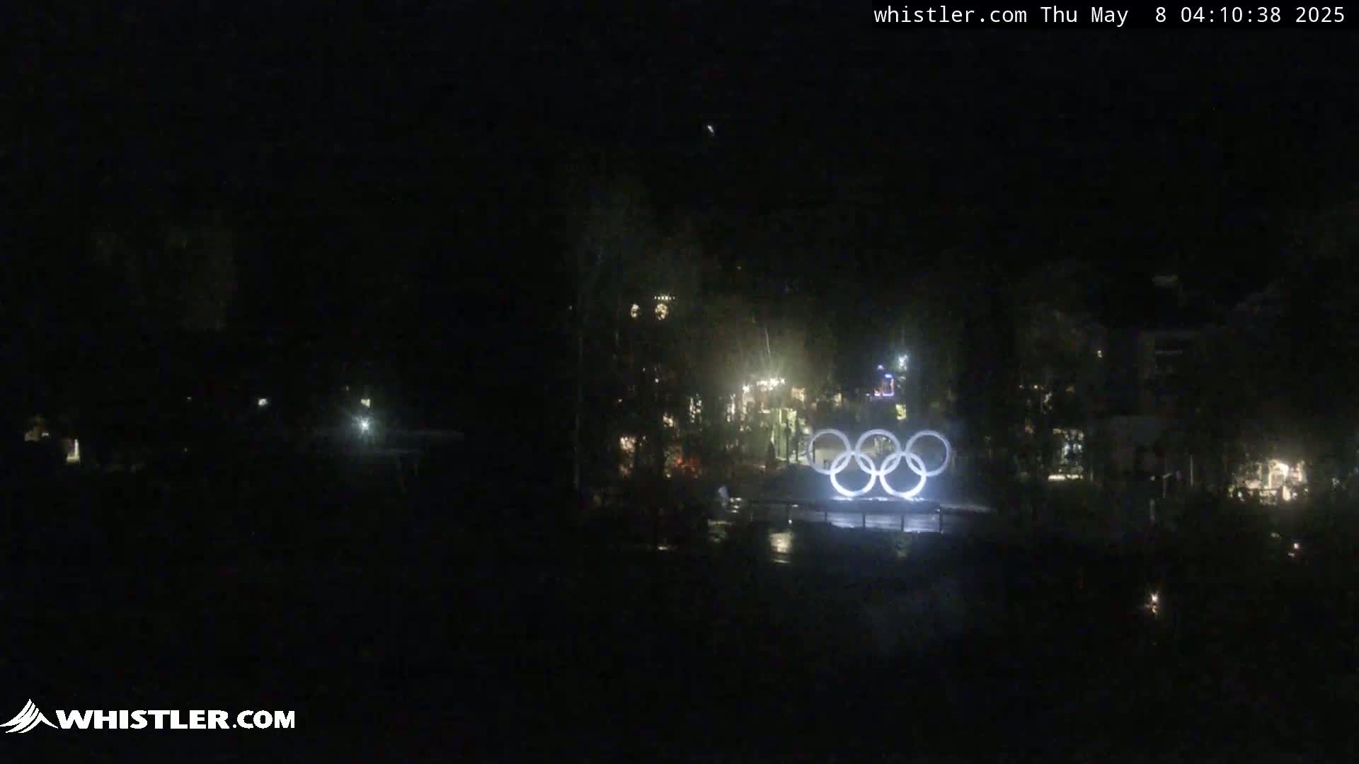 At night, illuminated Olympic rings are visible in the distance among dimly lit trees and buildings.