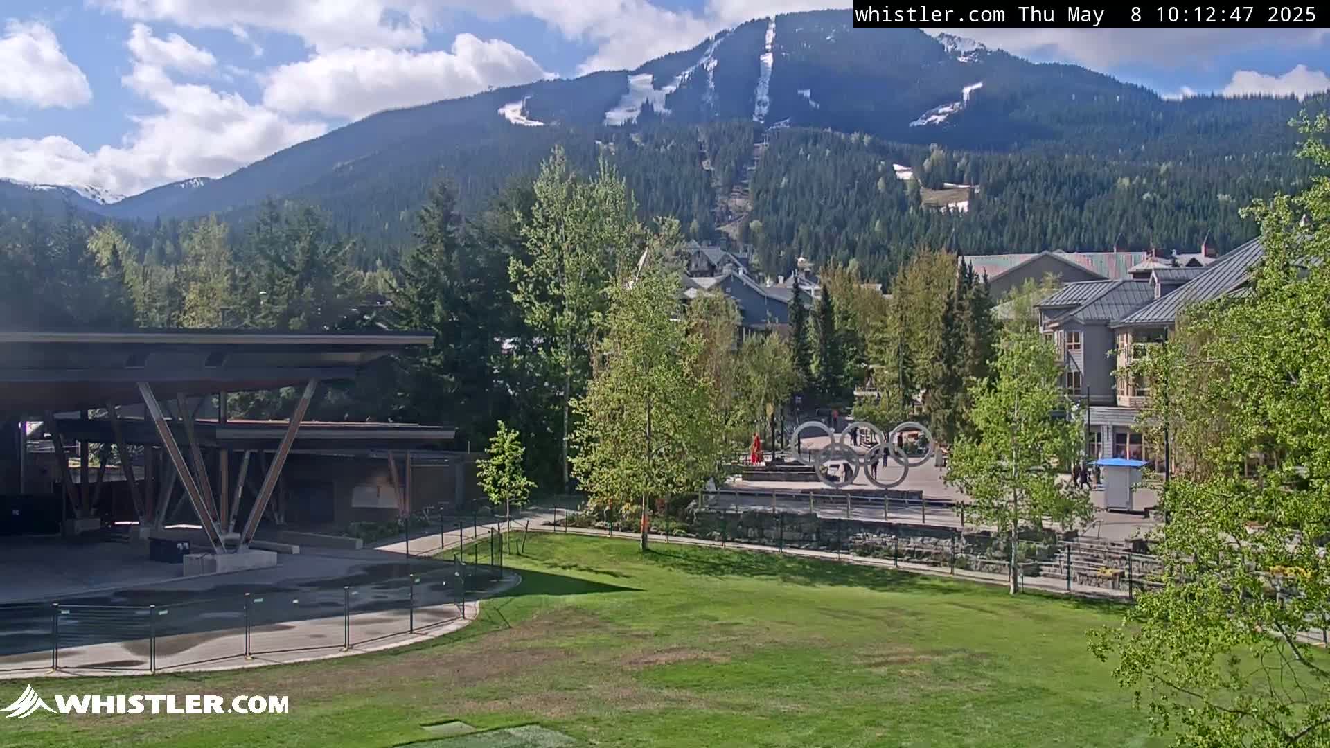 A partly sunny day reveals a mountain range with snow-capped peaks in the background, overlooking a village with buildings and green spaces, and a modern pavilion-like structure in the foreground.