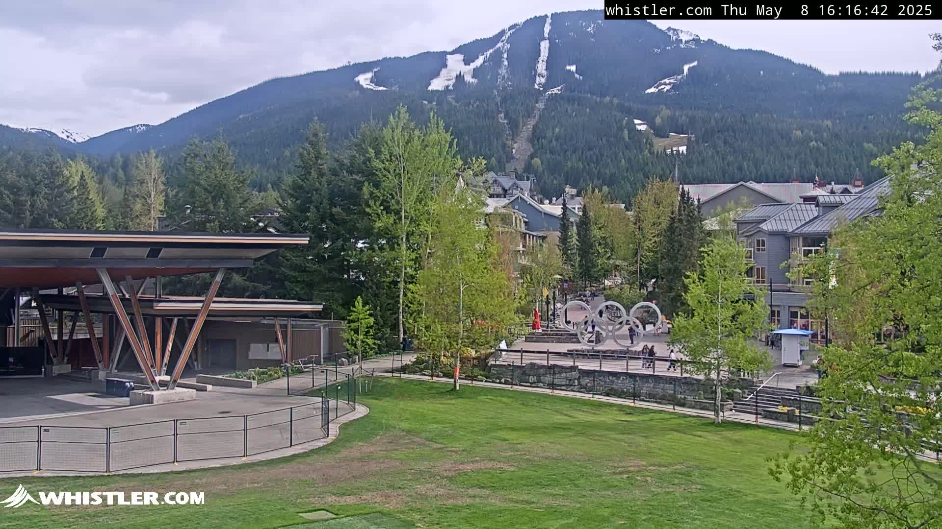 A partly cloudy day shows a mountainside ski resort with a village below, featuring a grassy area with a modern pavilion, Olympic rings, and several buildings.
