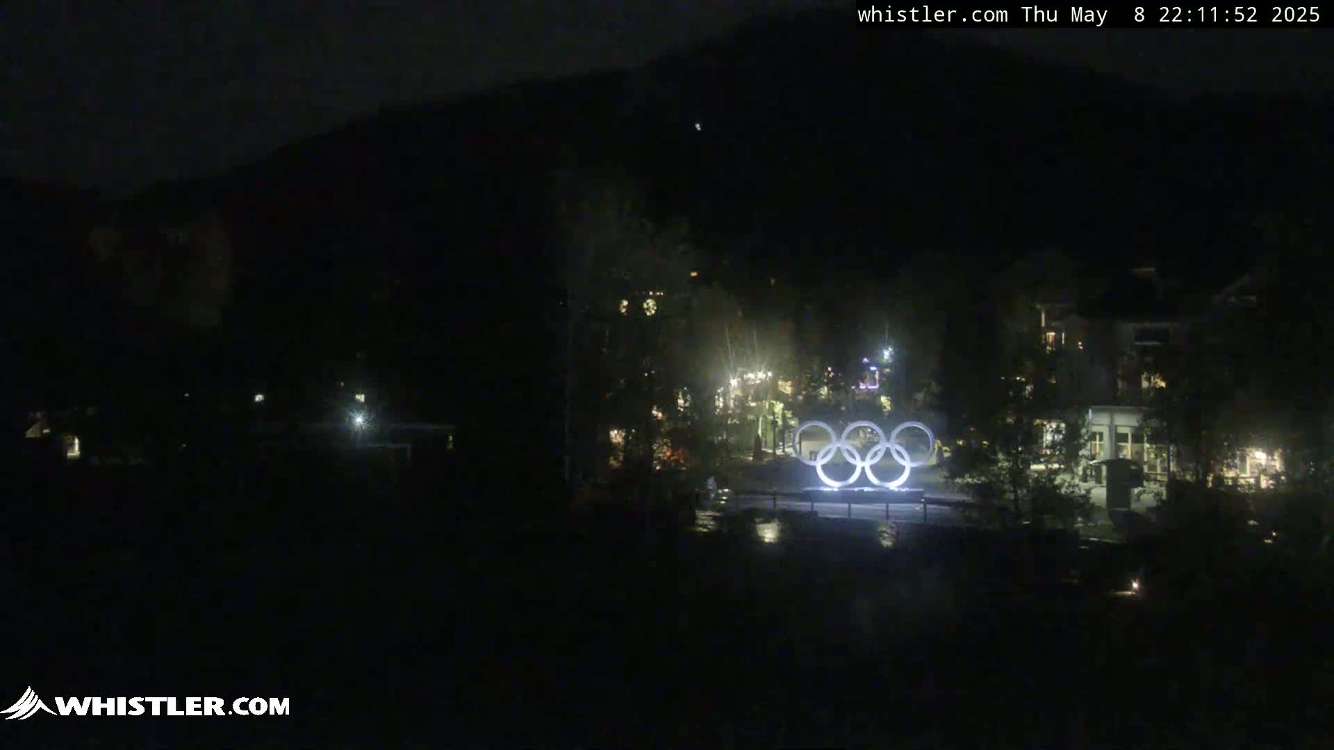 At night, illuminated Olympic rings are situated in front of a dark, shadowy village nestled against a mountainside.