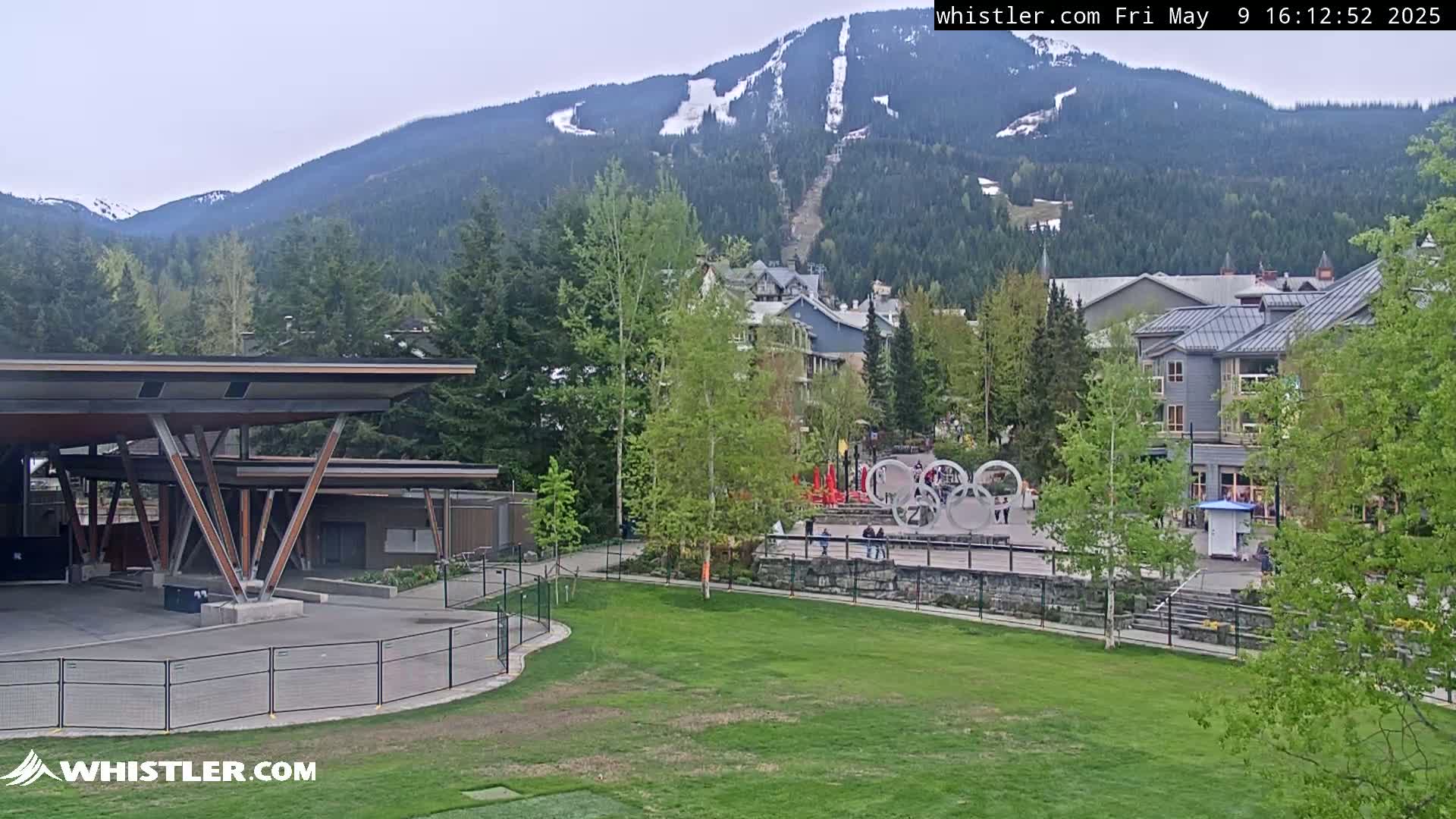 A partially cloudy day shows a mountain with a ski run, buildings, a grassy area with an outdoor stage and the Olympic rings in the foreground.