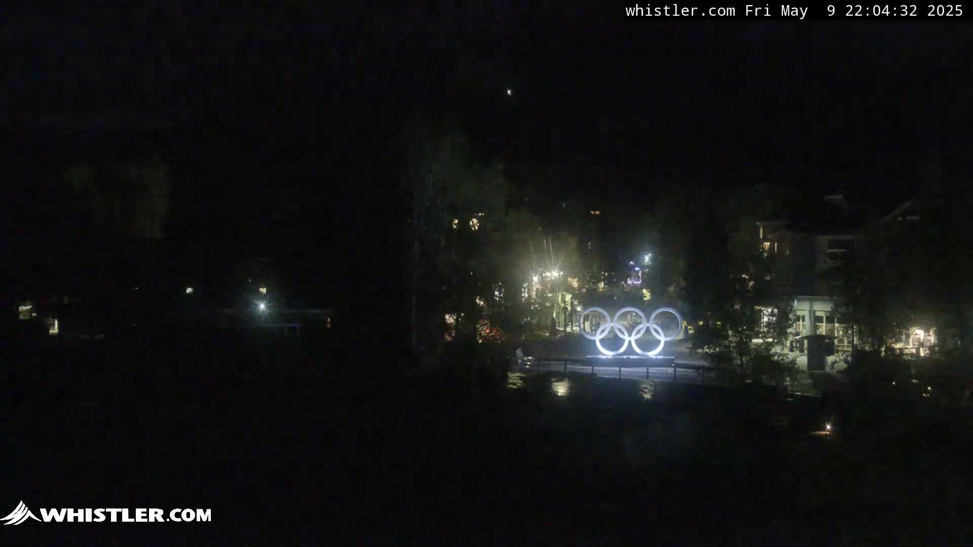At night, illuminated Olympic rings stand before a dark, sparsely lit village.