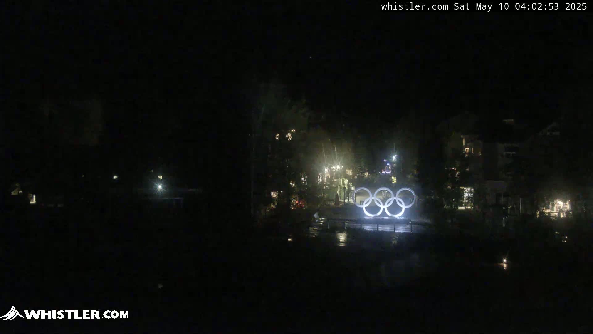 Illuminated Olympic rings stand in a dark, nighttime scene with a few scattered lights visible in the background.