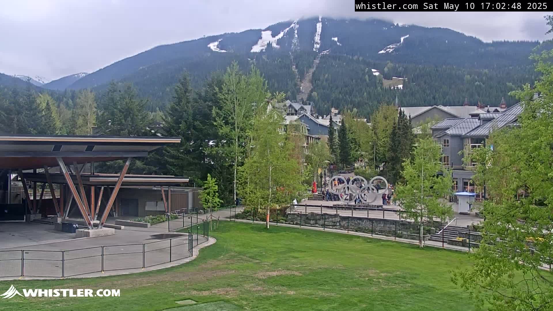 A partially cloudy day reveals a mountain village nestled in a valley with a grassy area in the foreground, featuring a modern structure and a large Olympic rings sculpture.