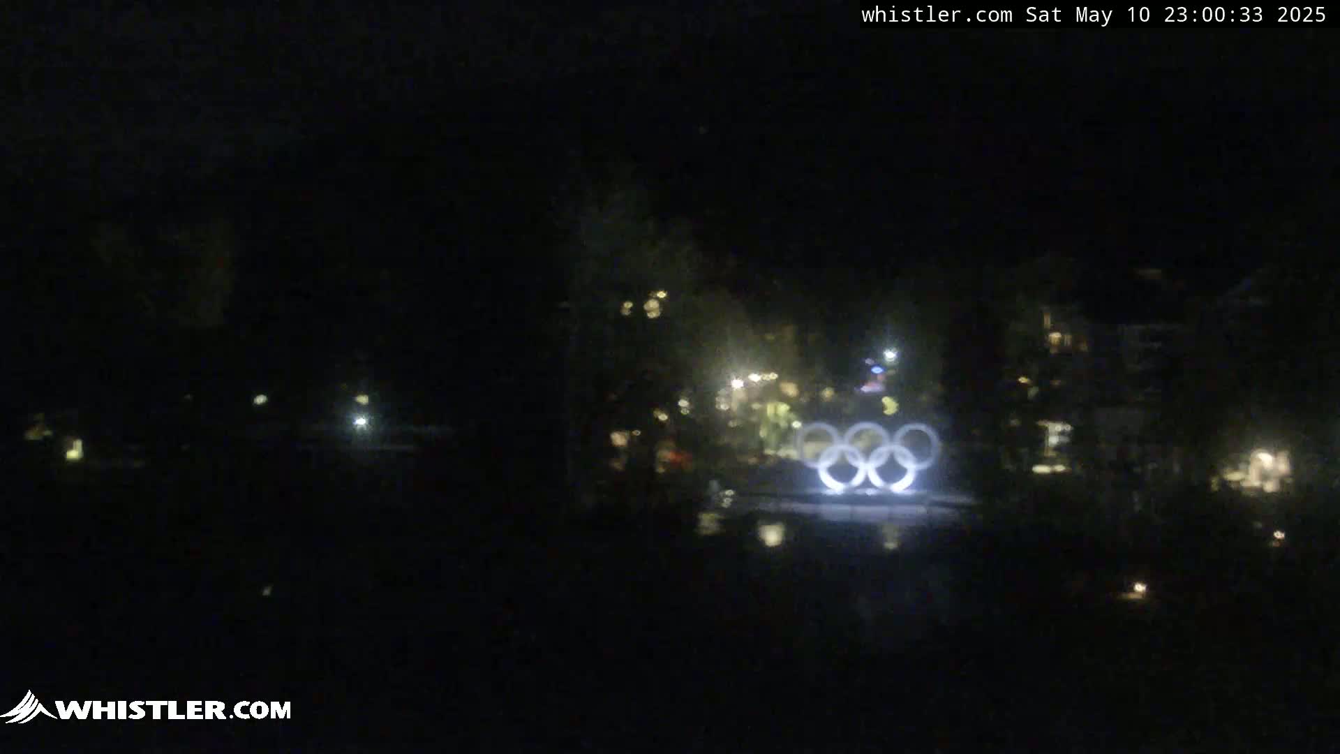 At night, illuminated Olympic rings are visible in the distance, with a few scattered lights in the dark surroundings.