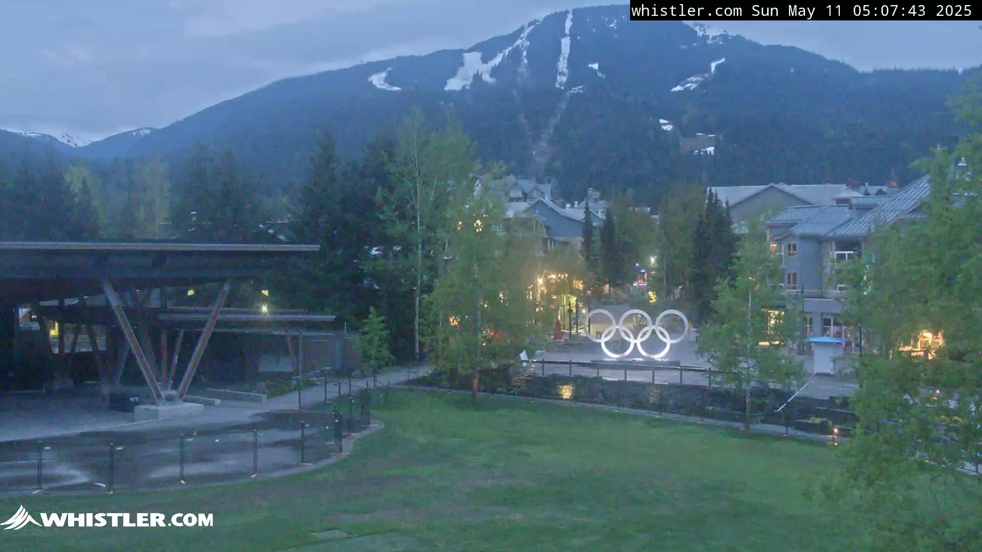 A view of a town nestled in a valley under a twilight sky, with mountains showing snow patches in the background and the Olympic rings visible in the foreground.