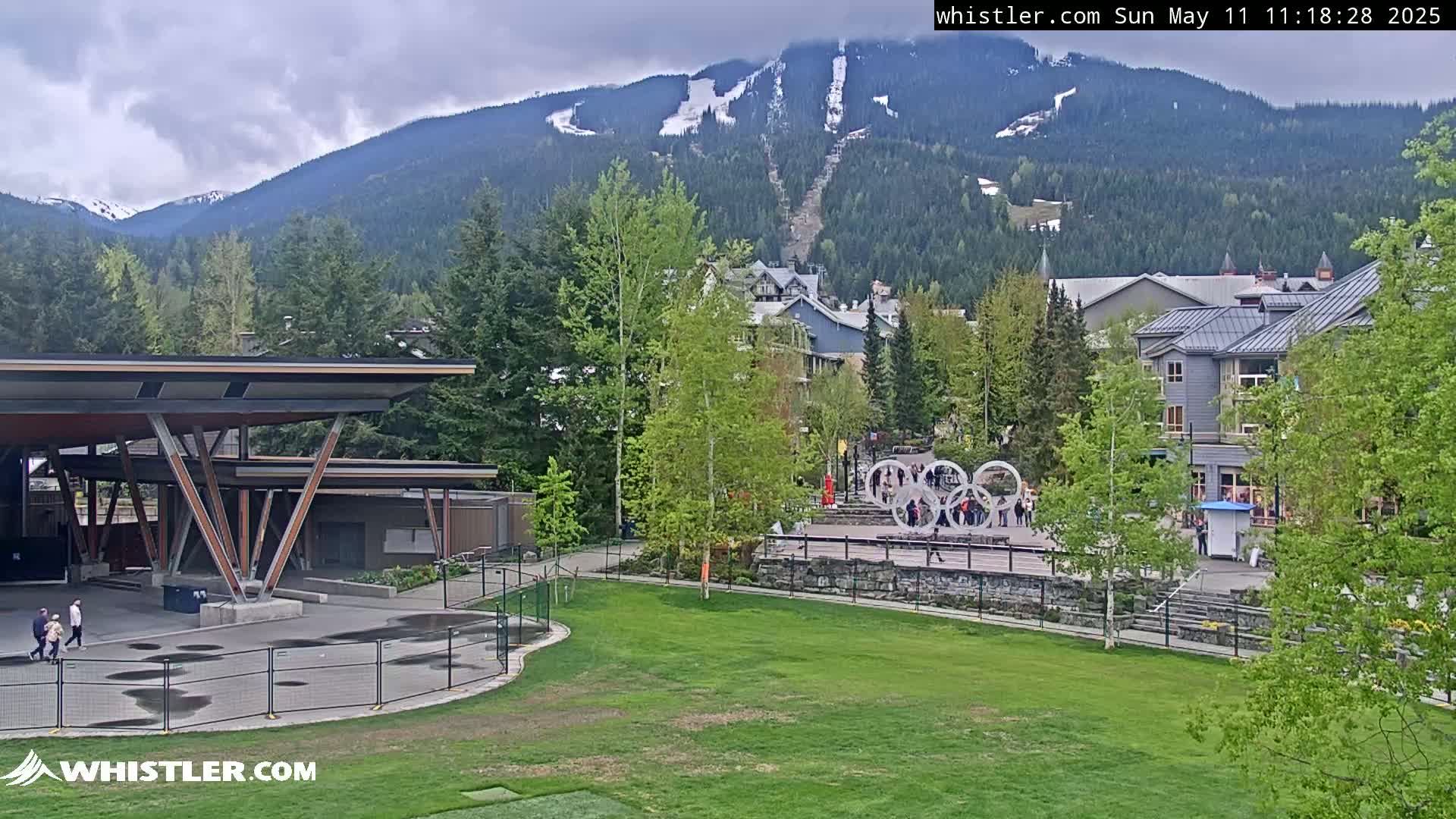 A grassy park with a covered pavilion and a few people, situated in front of buildings and trees, with snow-capped mountains in the background under overcast skies.