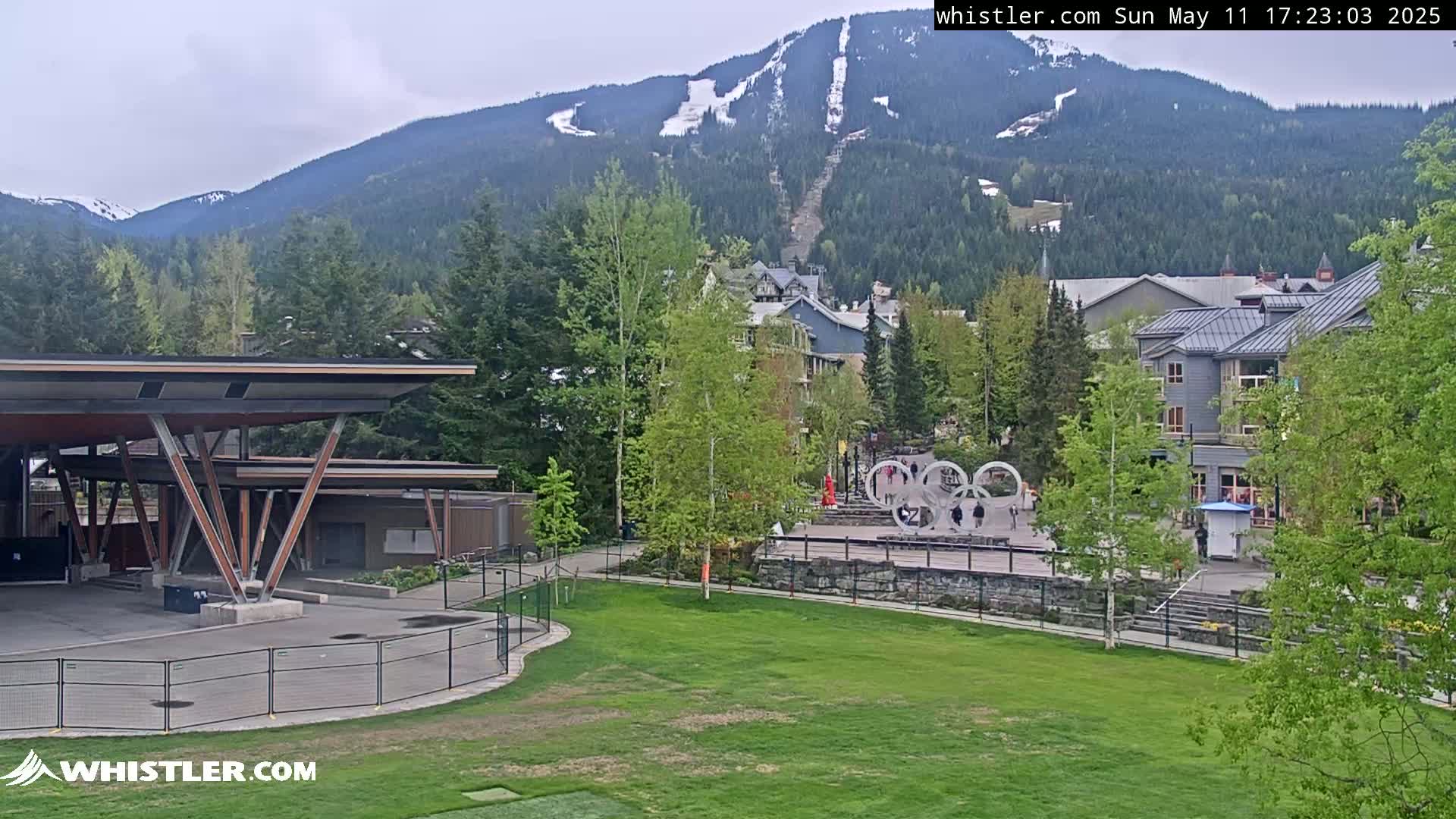 A grassy area with a modern structure and buildings sits in front of a mountain range with some remaining snow, under an overcast sky.