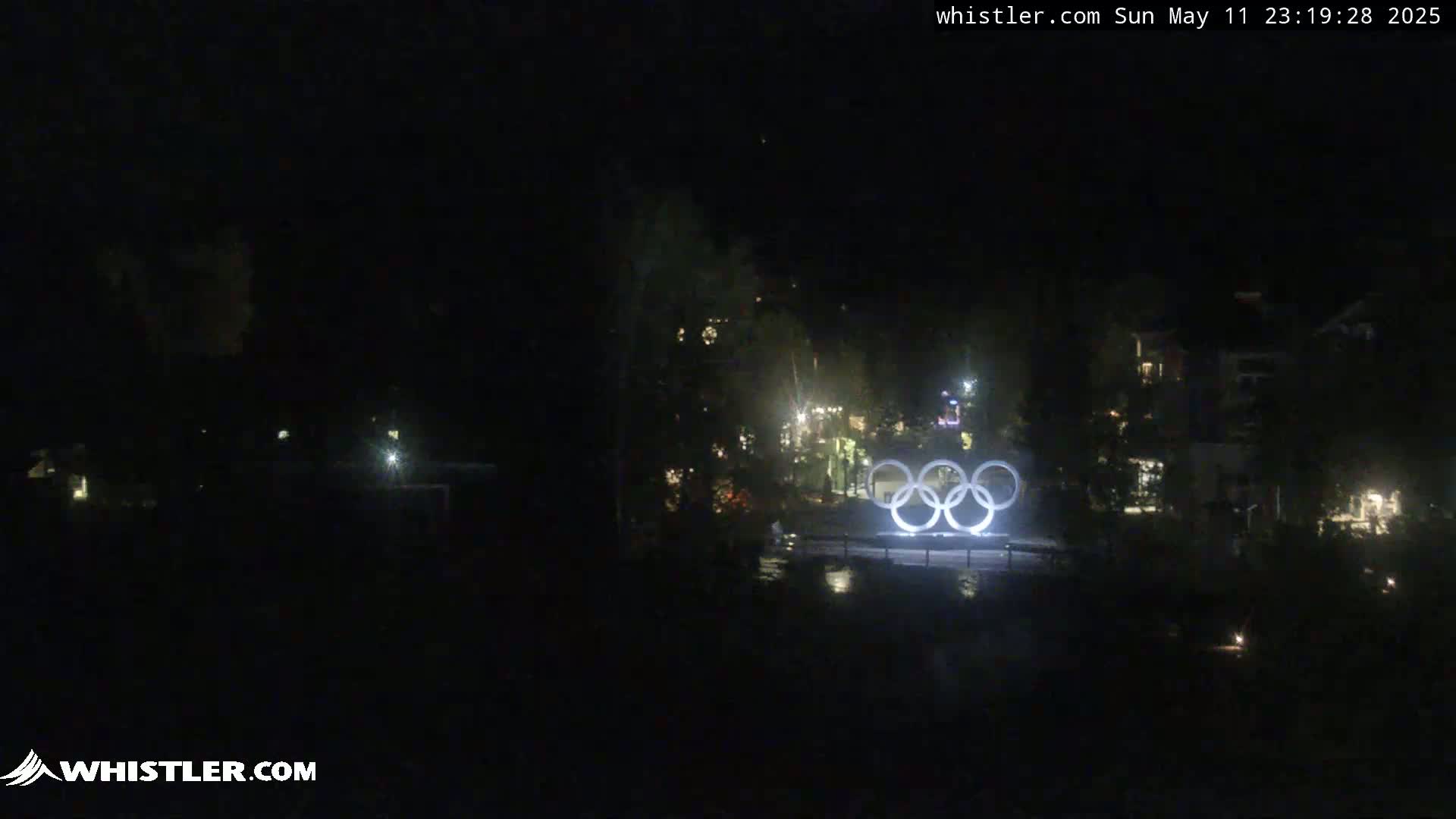 At night, the illuminated Olympic rings are visible in a dark, mostly obscured area with a few lights scattered in the background.