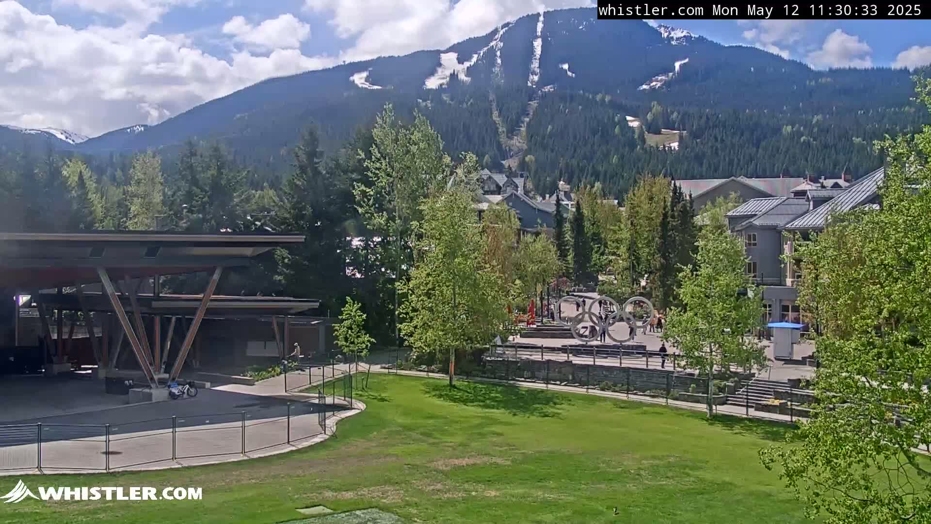 A sunny view of a mountain village shows a grassy area with a modern structure, buildings, and the Olympic rings sculpture in the foreground, with mountains and a ski resort visible in the background.