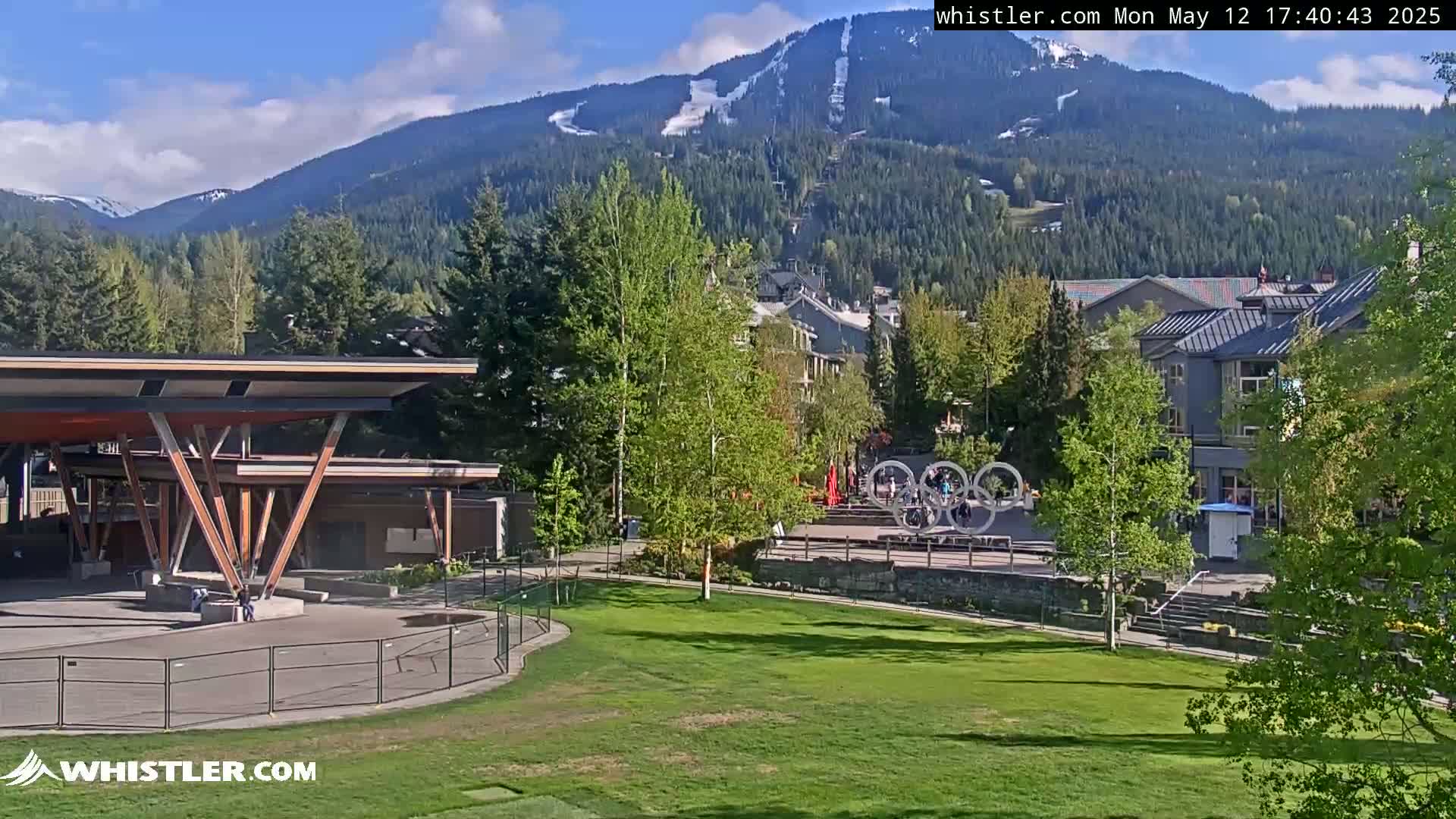 A sunny view of a grassy park area with a modern pavilion, buildings, and a snow-capped mountain range in the background.