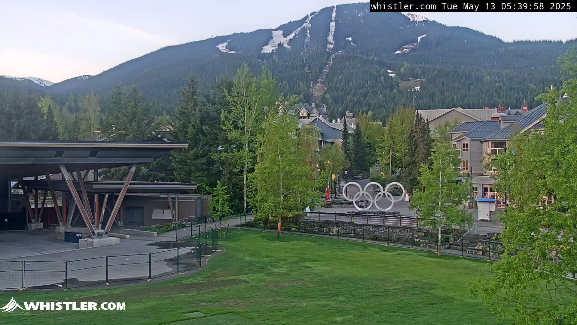 A grassy field overlooks an outdoor stage, buildings, and the Olympic rings, all nestled in a valley against a backdrop of snow-capped mountains under a mostly clear sky.