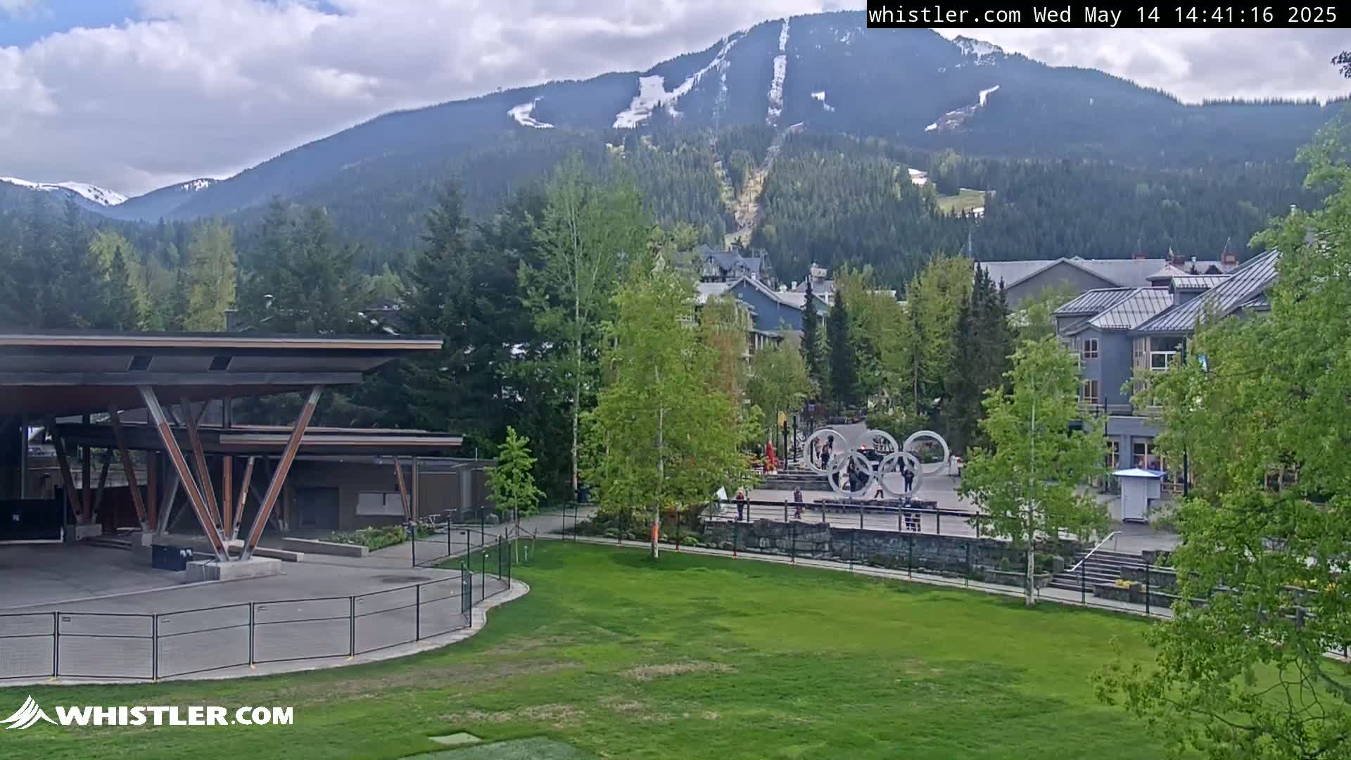 A partly cloudy day shows a mountain village with a grassy park, a modern pavilion, and snow-capped mountains in the background.